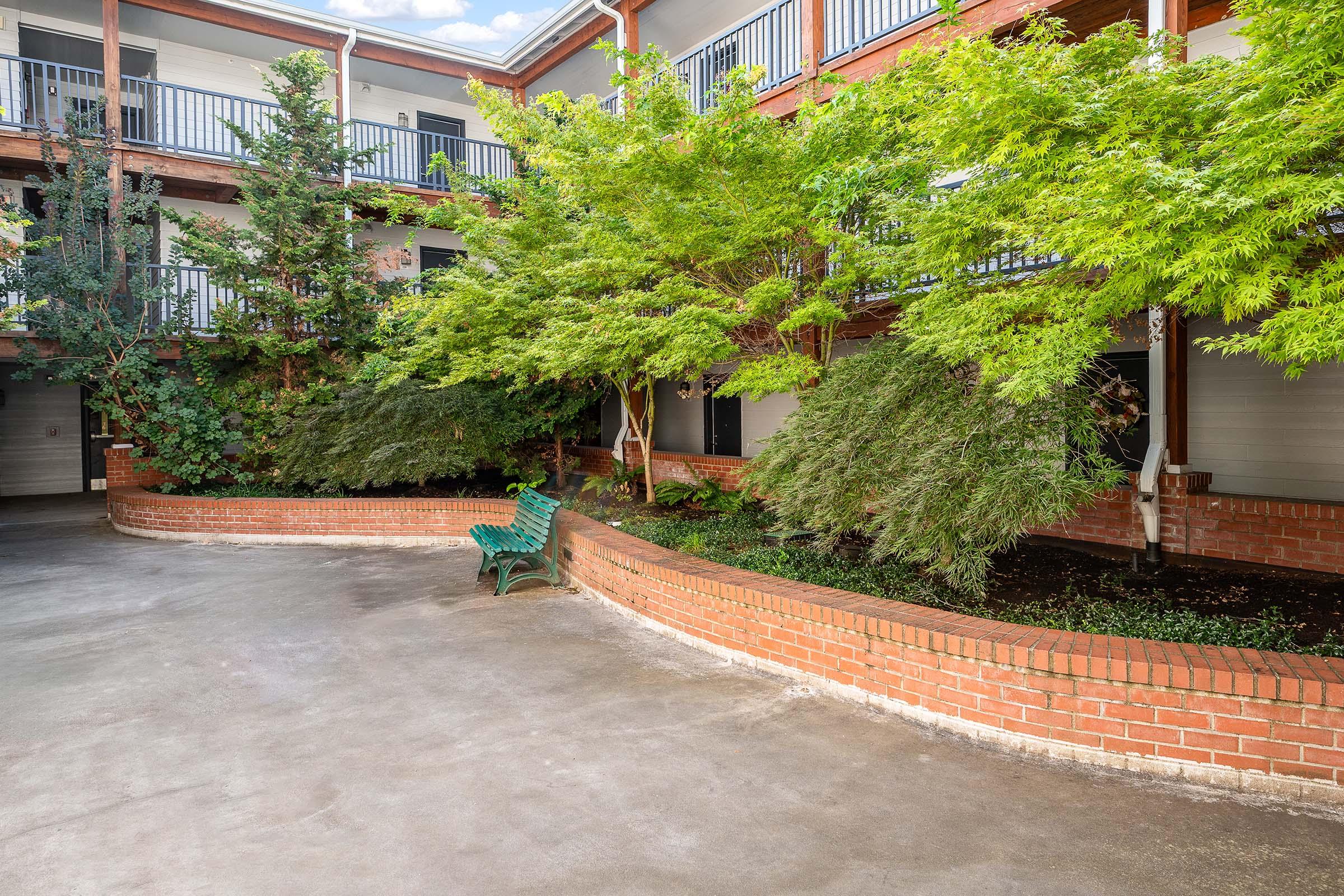 A peaceful courtyard with green foliage and trees, surrounded by brick walls. A curved, brick planter contains shrubs, and a green bench is situated nearby. The area features multiple levels of flooring, with balconies visible above, creating a serene atmosphere.