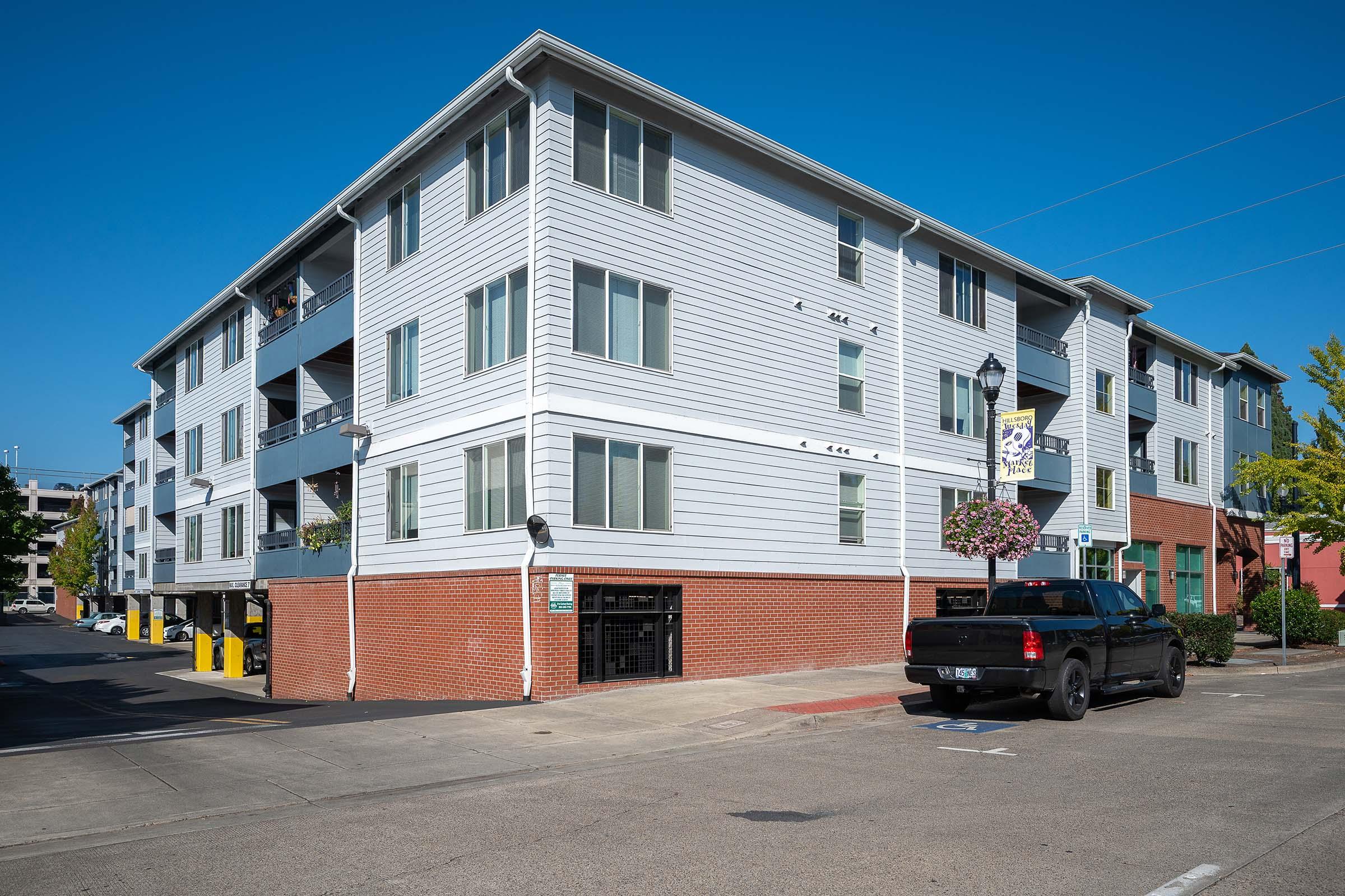 A modern, multi-story apartment building with a light gray exterior and several windows. The building features a brick base and a parking lot in front, where a black pickup truck is parked. Green trees and flowering plants add a touch of greenery to the scene under a clear blue sky.