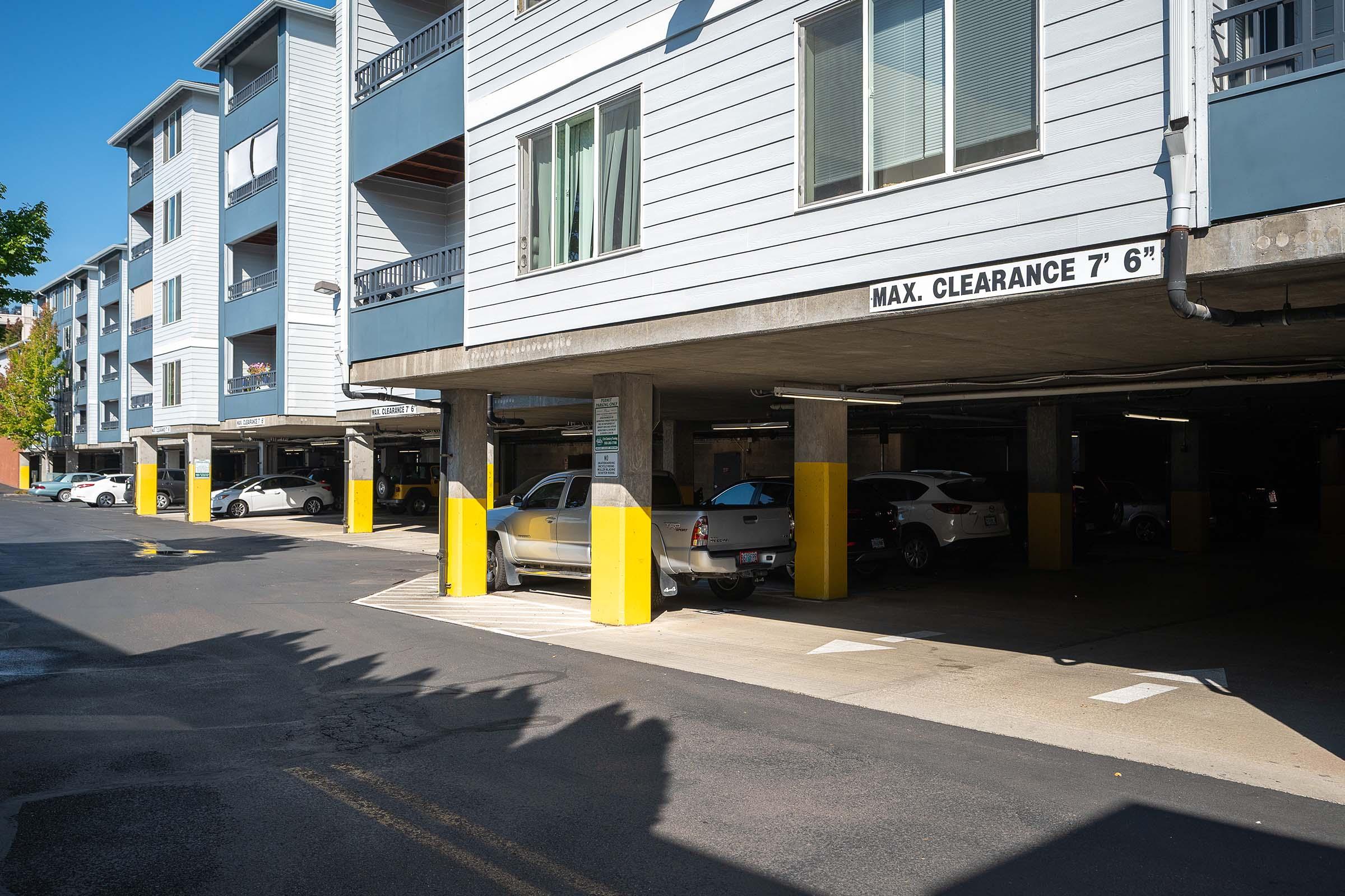 View of an underground parking garage with several parked cars. The structure features yellow-painted pillars and a sign indicating the maximum clearance of 7 feet 6 inches. Sunlight casts shadows across the pavement, and the layout includes designated parking spaces.
