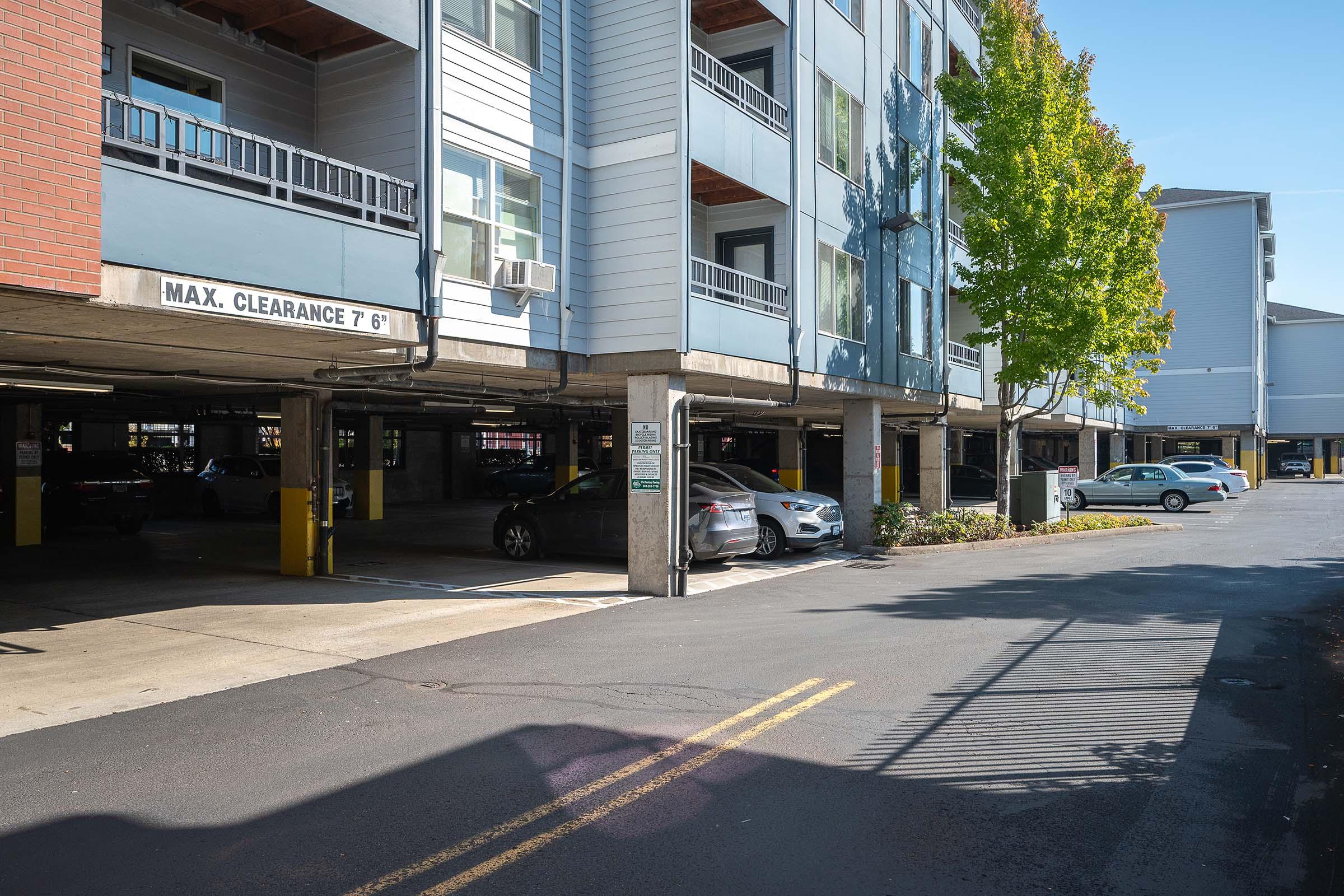 View of an outdoor parking area featuring several parked cars beneath a multi-story residential building. Concrete support pillars, signage indicating maximum clearance of 7 feet, and well-maintained surrounding landscape are visible. Bright sunlight illuminates the scene.
