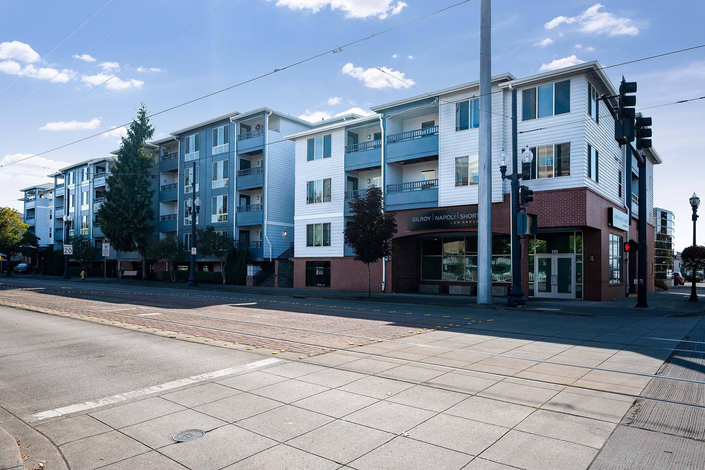 A street view of a multi-story apartment building featuring a mix of blue and white siding. The ground level has a storefront or office space with large windows, while traffic lights and nearby trees are visible. Smooth pavement lines the area in front, indicating a well-maintained urban environment.
