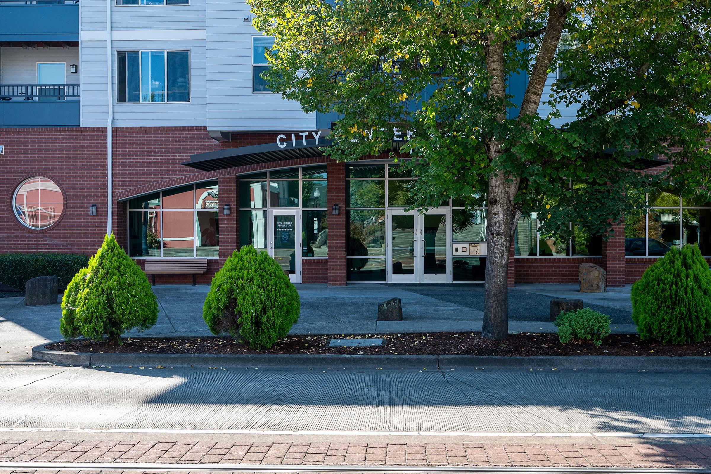 A city building entrance featuring large glass doors, surrounded by neatly trimmed green bushes and trees. The architecture includes modern elements with brick and siding. Sidewalk and street visible in the foreground, with a clear blue sky above.