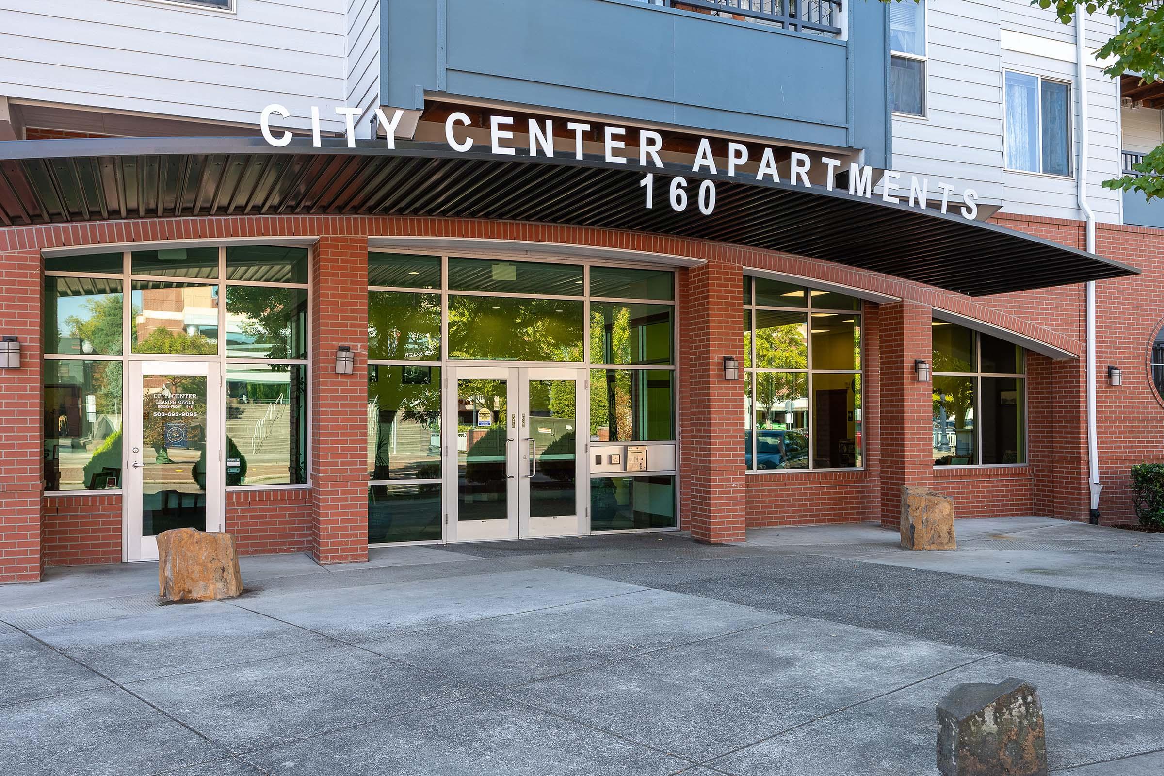Entrance to City Center Apartments, featuring large glass doors and windows, a modern façade with brick accents, and a canopy overhead. The building is well-lit, surrounded by greenery, and displays the address "160" prominently above the entrance.
