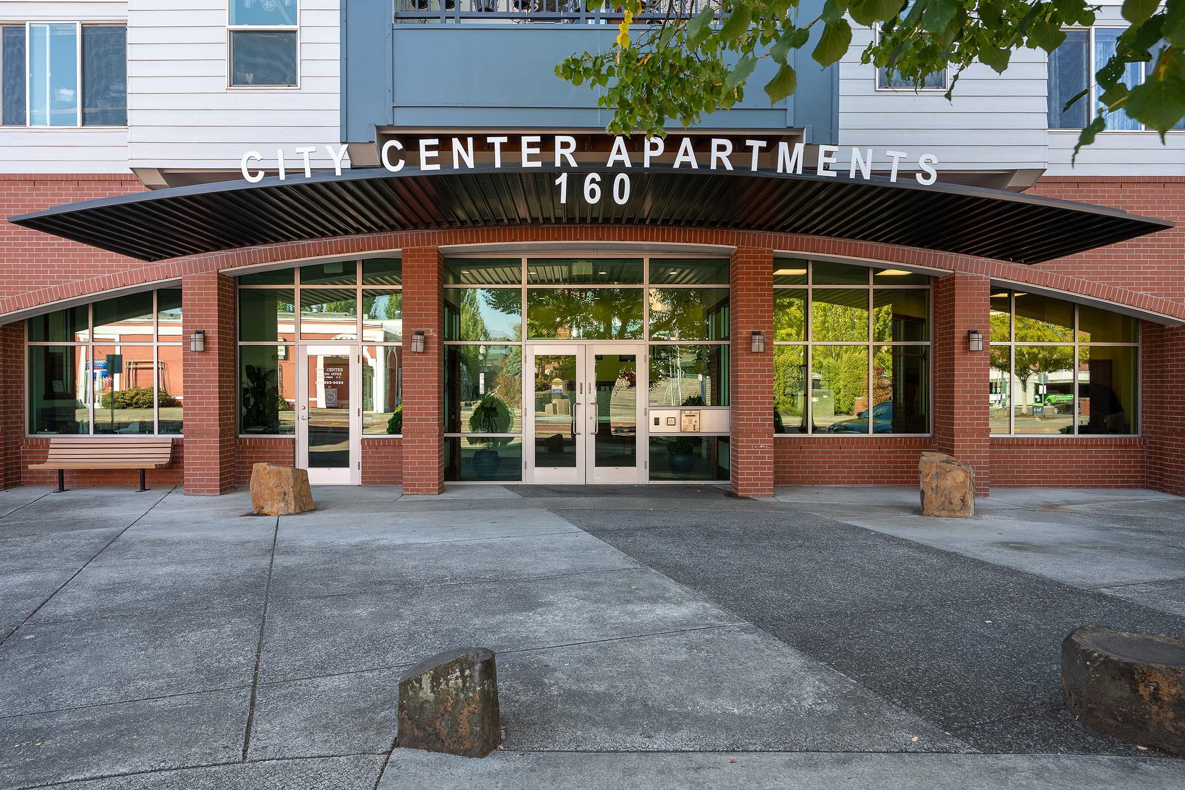 Entrance of City Center Apartments, featuring large glass doors under an overhang, with the building name prominently displayed above. Surrounding the entrance are stone benches and landscaped greenery, set in a modern urban environment.
