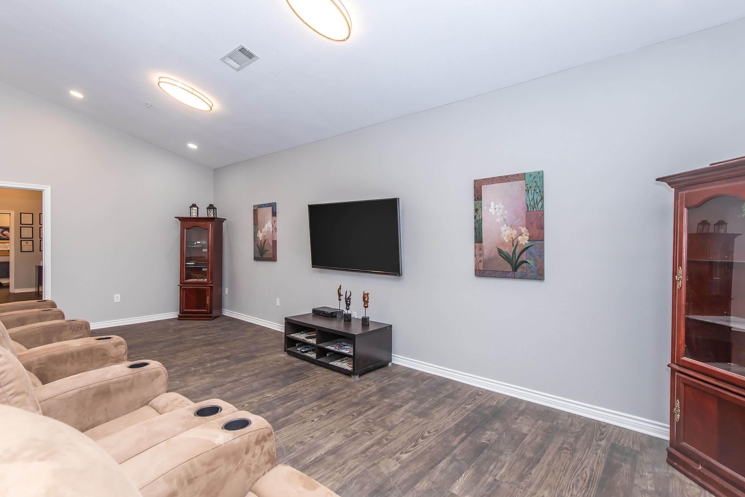 A bright, modern living room featuring a wall-mounted flat-screen TV, two decorative paintings, and a wooden cabinet. Comfortable beige recliners are arranged for seating, and the flooring is a rich, dark wood. The walls are painted in a light gray color, creating a serene atmosphere.