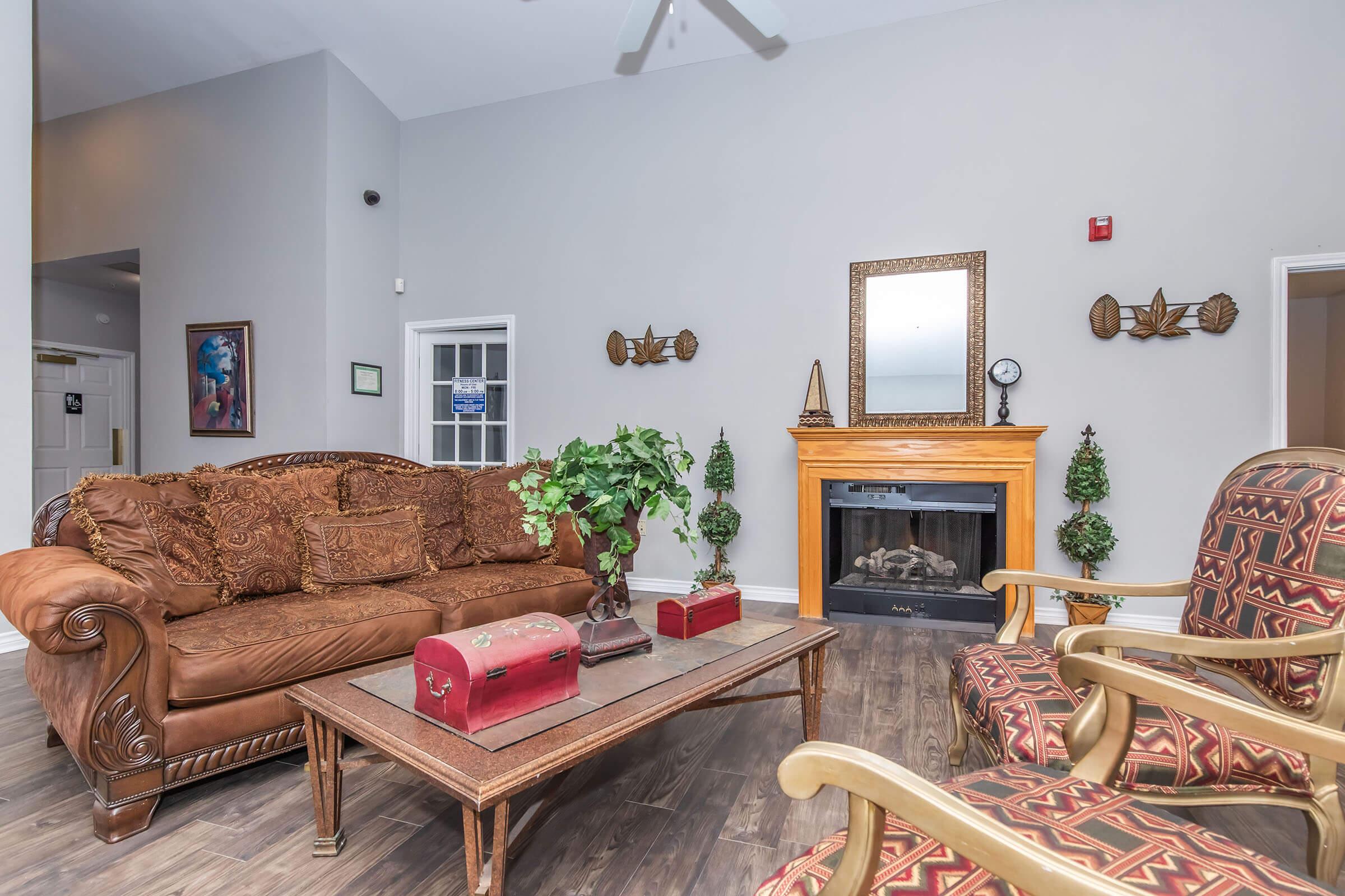 A cozy living room with a brown sofa and decorative pillows, a wooden coffee table with a red chest, and two patterned armchairs. The space features a fireplace with a wooden mantel, framed mirrors on the walls, and potted greenery, creating a warm and inviting atmosphere.