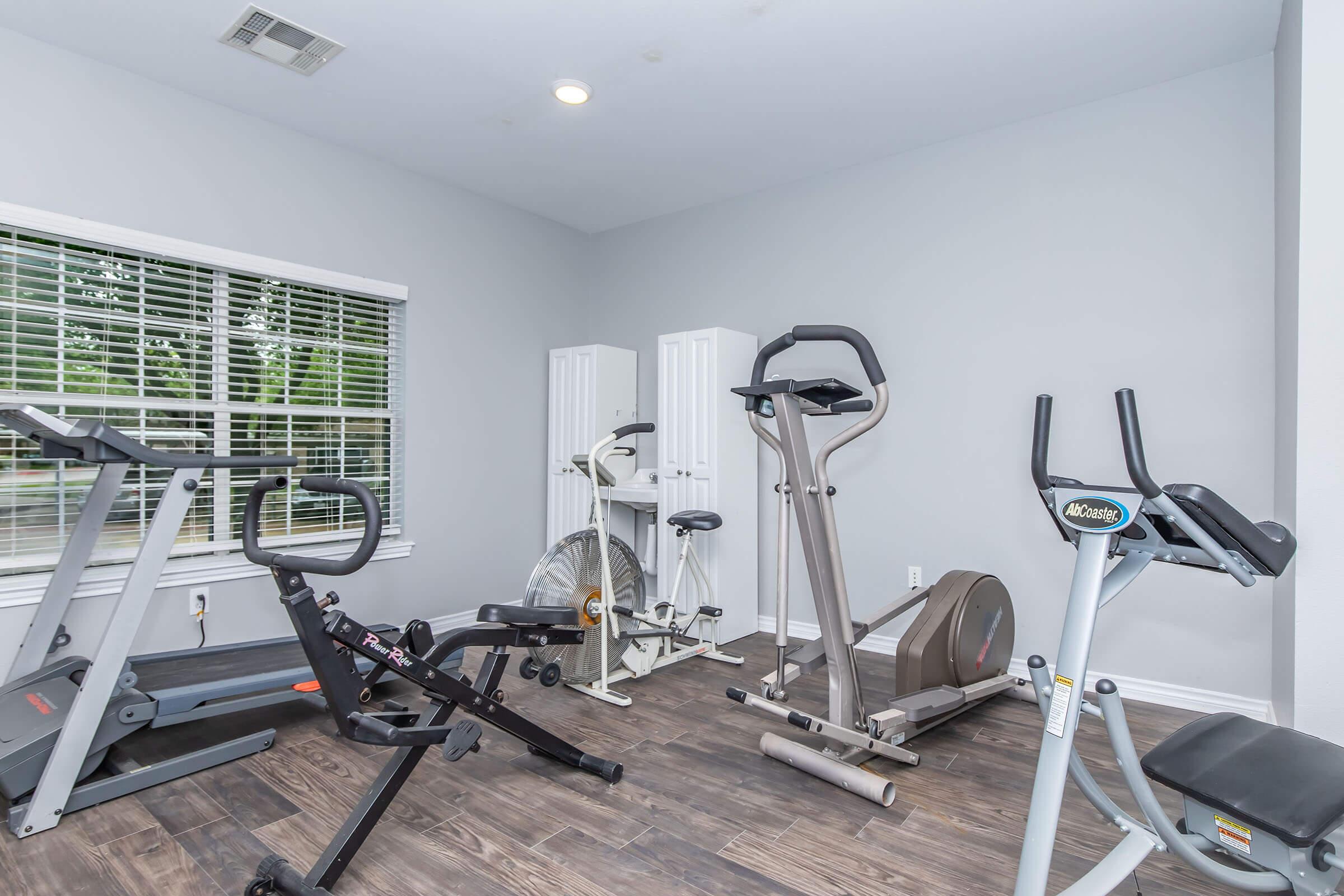 A small home gym with various exercise equipment, including treadmills, an elliptical machine, a stationary bike, and a fan bike. The room features light gray walls and large windows with blinds, allowing natural light to enter, and a wood-like floor.