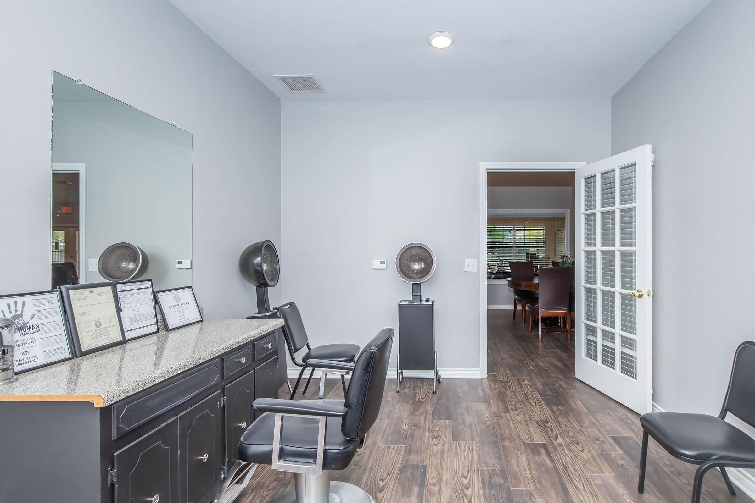 Interior of a hair salon featuring a grey wall, a granite countertop with salon certificates displayed, two black salon chairs, and hair styling equipment. A doorway leads to another room, which appears to have a dining area in view. Natural light enters the space, creating a bright atmosphere.