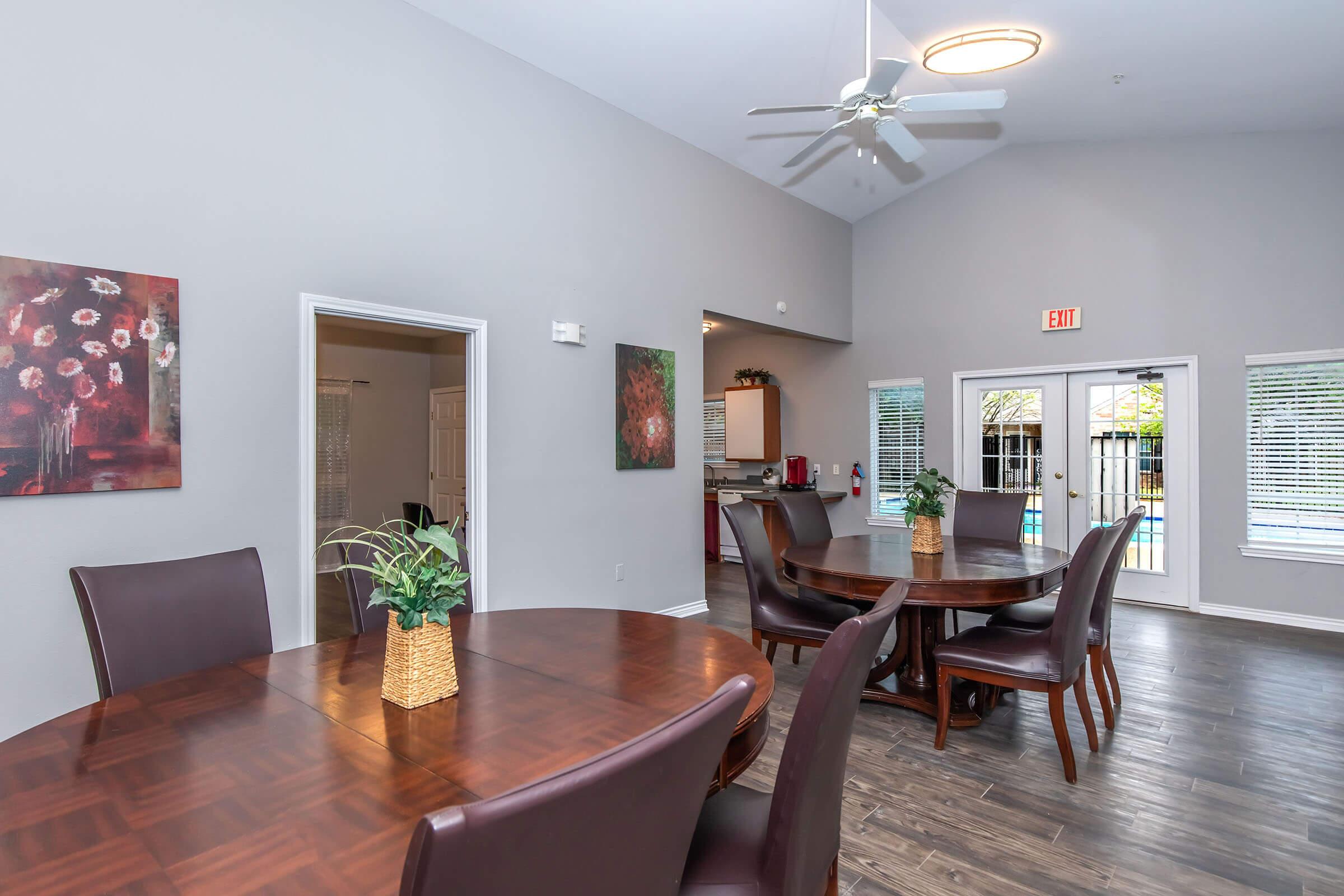 A well-lit community space featuring dark wooden tables with brown leather chairs. There are decorative flower paintings on the walls, and a large window showing a view of an outdoor pool area. A ceiling fan hangs above, contributing to a modern and inviting atmosphere.