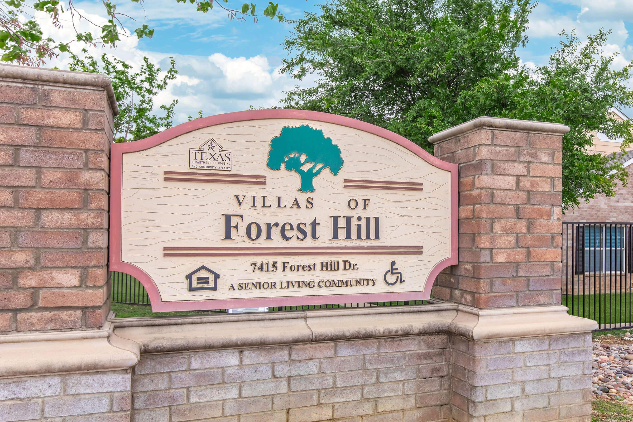 Sign for "Villas of Forest Hill," located at 7415 Forest Hill Dr. The sign features a tree icon and indicates that it is a senior living community, with accessibility symbols. The background includes a brick structure and green foliage.
