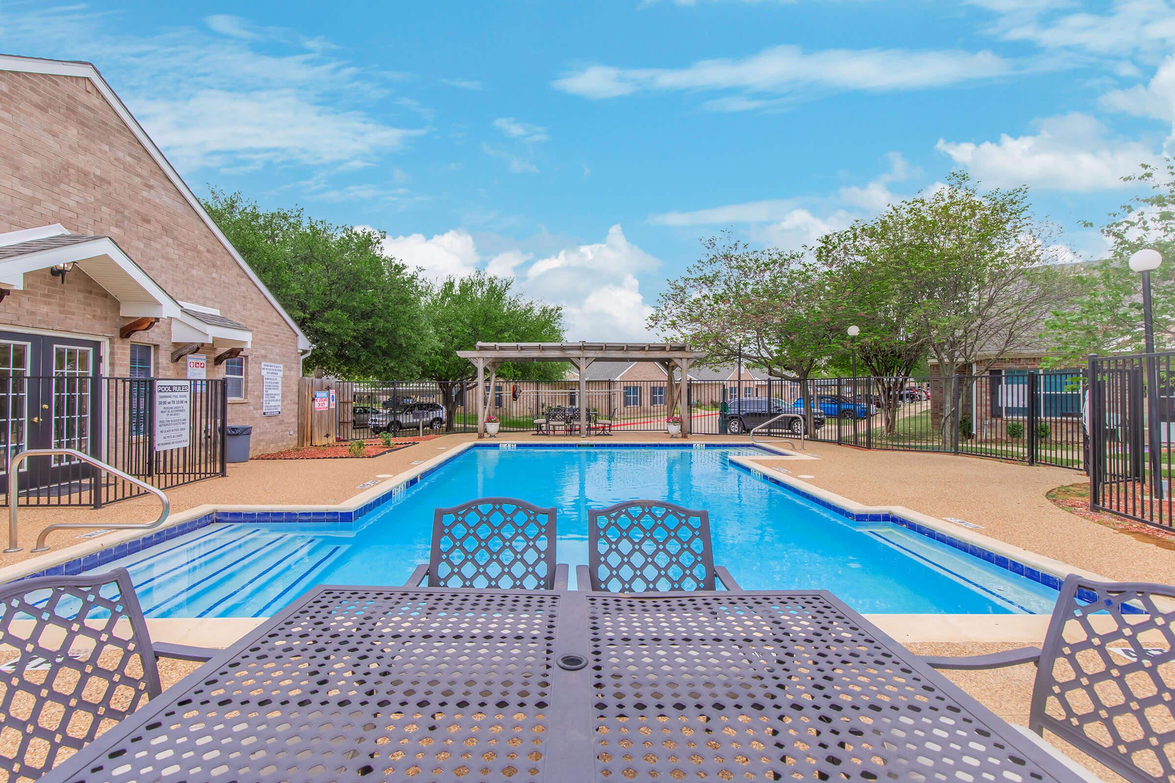 A clear blue swimming pool surrounded by lounge chairs and shaded by trees. Two buildings with light-colored brick exteriors are visible in the background, along with a pergola and additional seating areas. The sky is bright and partly cloudy, creating a relaxing outdoor atmosphere.