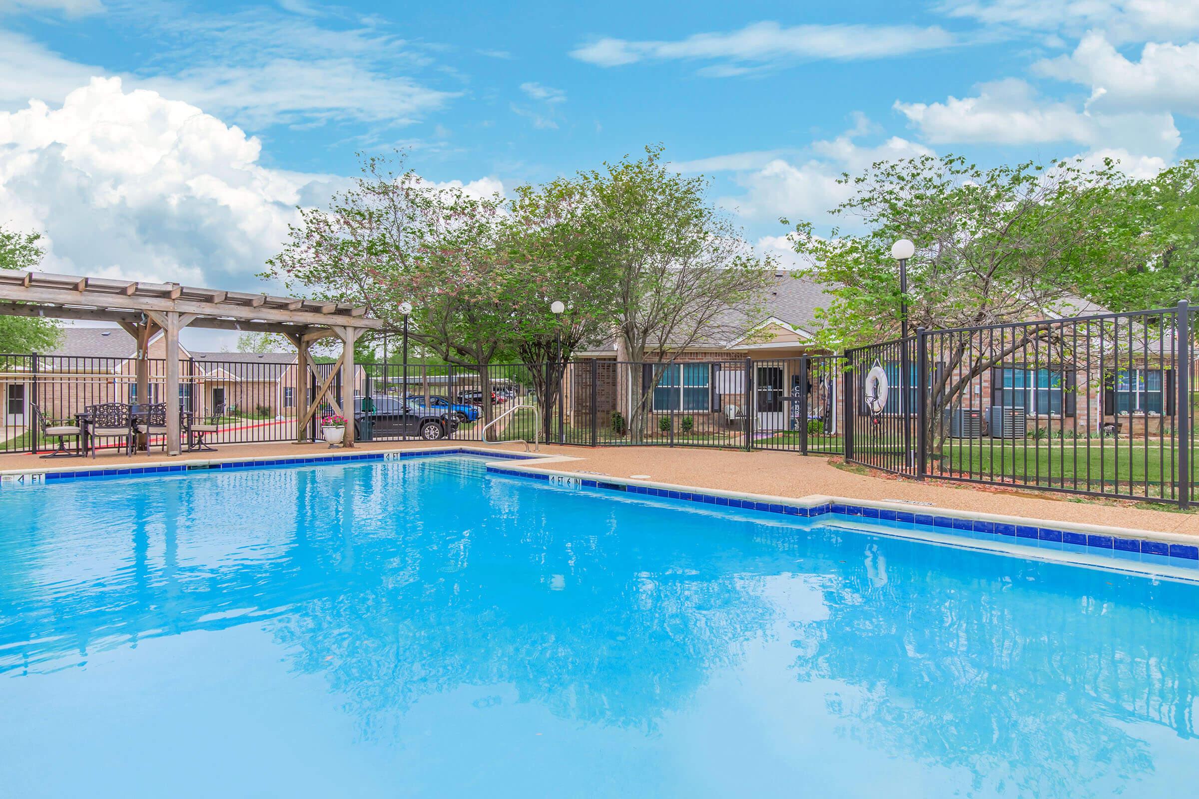 A bright outdoor swimming pool surrounded by lush greenery and a wooden pergola. The water is crystal clear, reflecting the blue sky and fluffy clouds above. Nearby, there are chairs and a fence adding to the inviting atmosphere of a relaxing outdoor space.