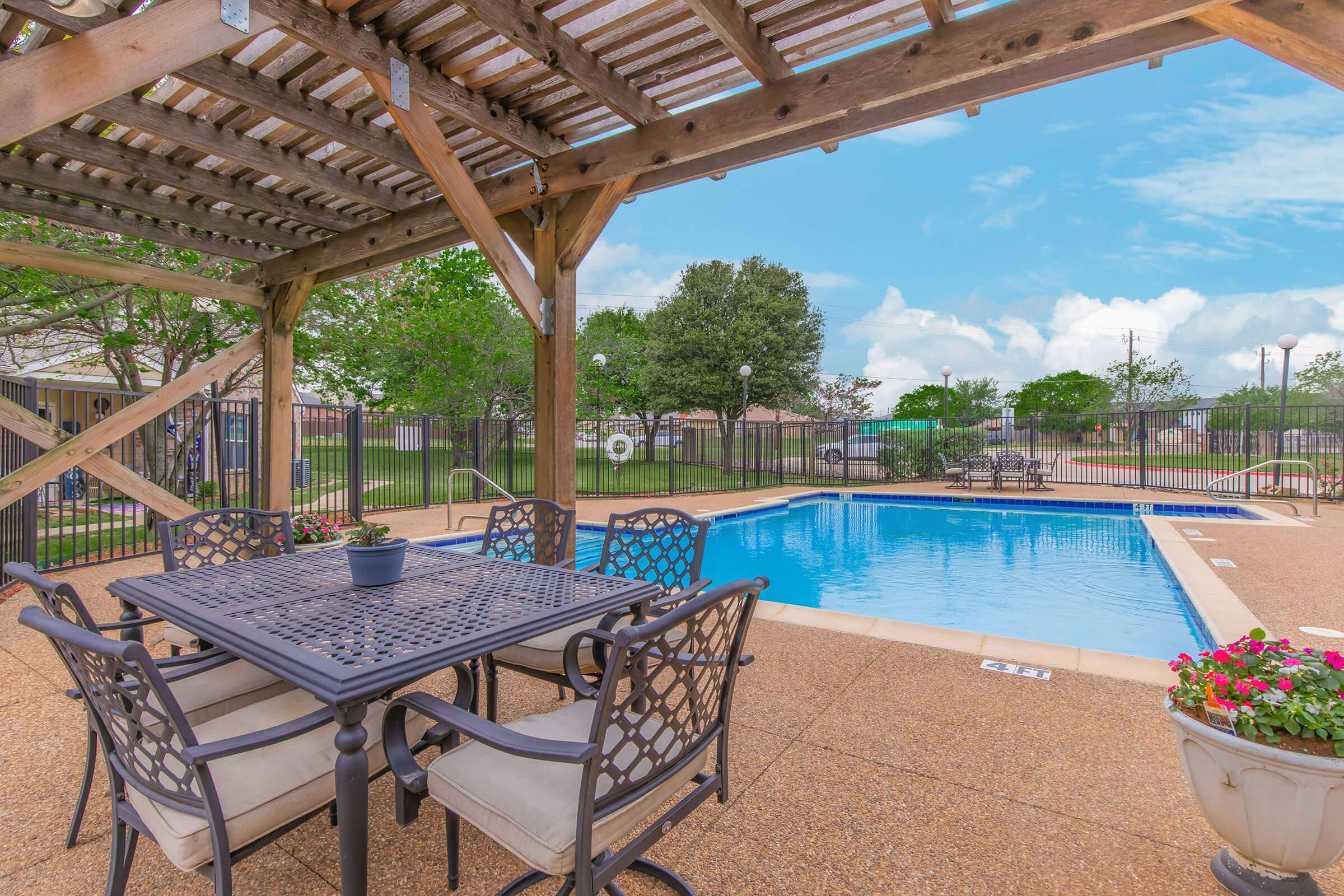 A shaded outdoor area featuring a dining table and chairs near a swimming pool. The pool is surrounded by a fence and includes lounge chairs. Lush green trees and shrubs are visible in the background, with a clear blue sky above. Flower pots add color to the scene.