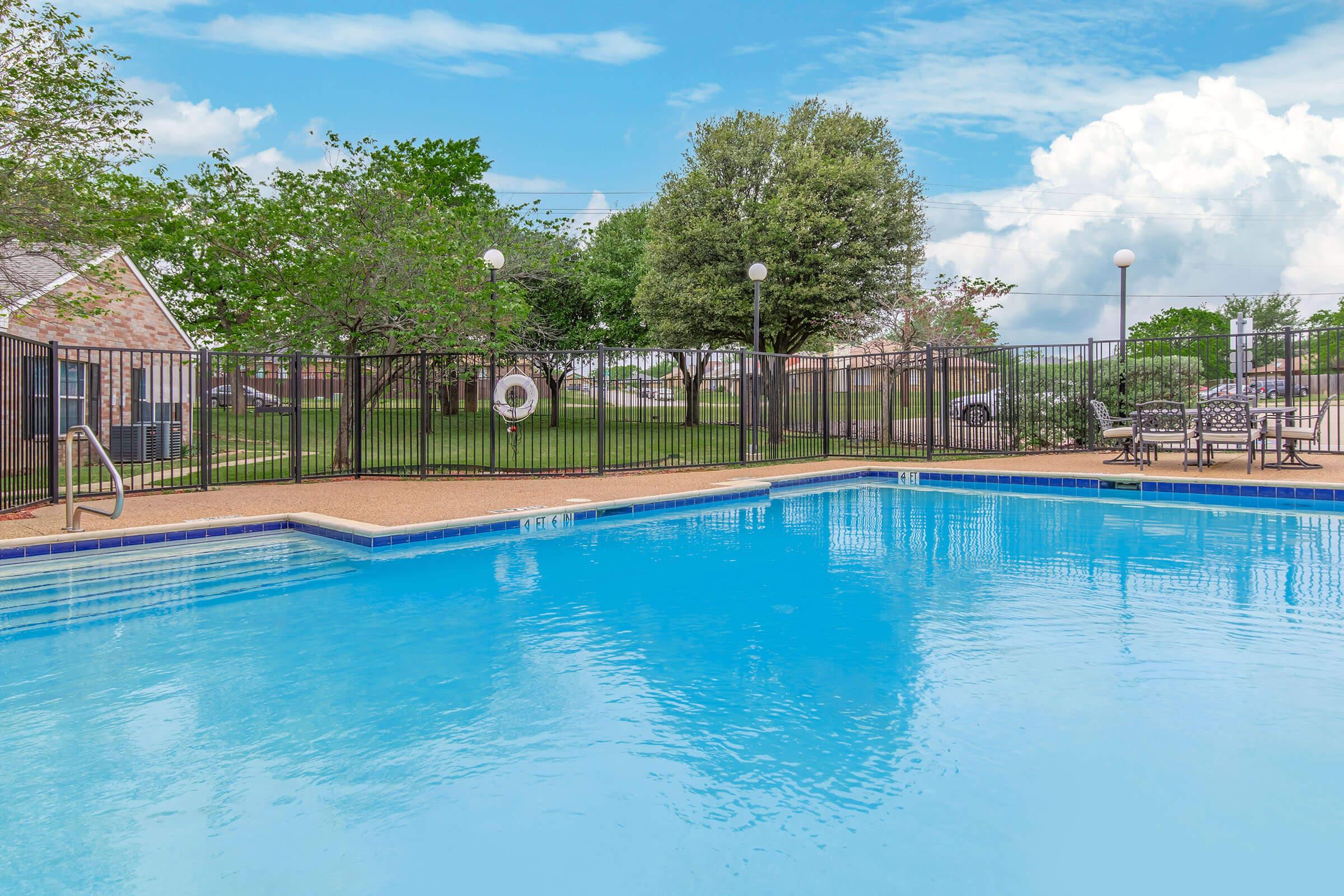 A clear blue swimming pool surrounded by a fenced area. In the background, there are lush green trees and grass, with a few tables and chairs nearby. The sky is partly cloudy, creating a bright and inviting atmosphere.