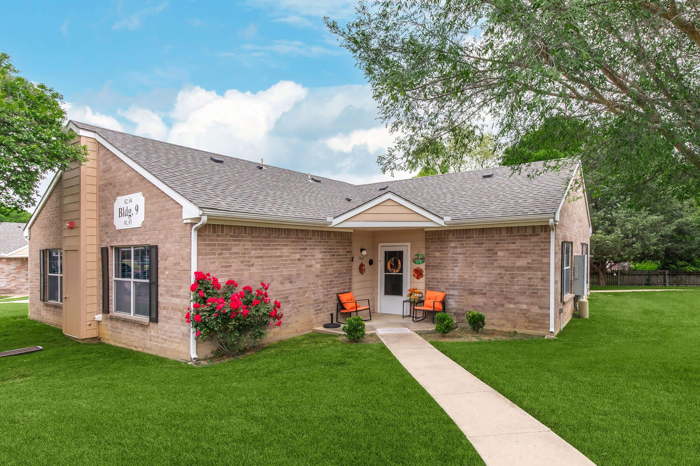 A single-story, brick exterior house with a well-manicured front lawn and flower beds. The entrance features a small porch with two orange chairs, and decorative elements like potted plants and a welcome sign. Lush green trees provide background foliage against a partly cloudy sky.