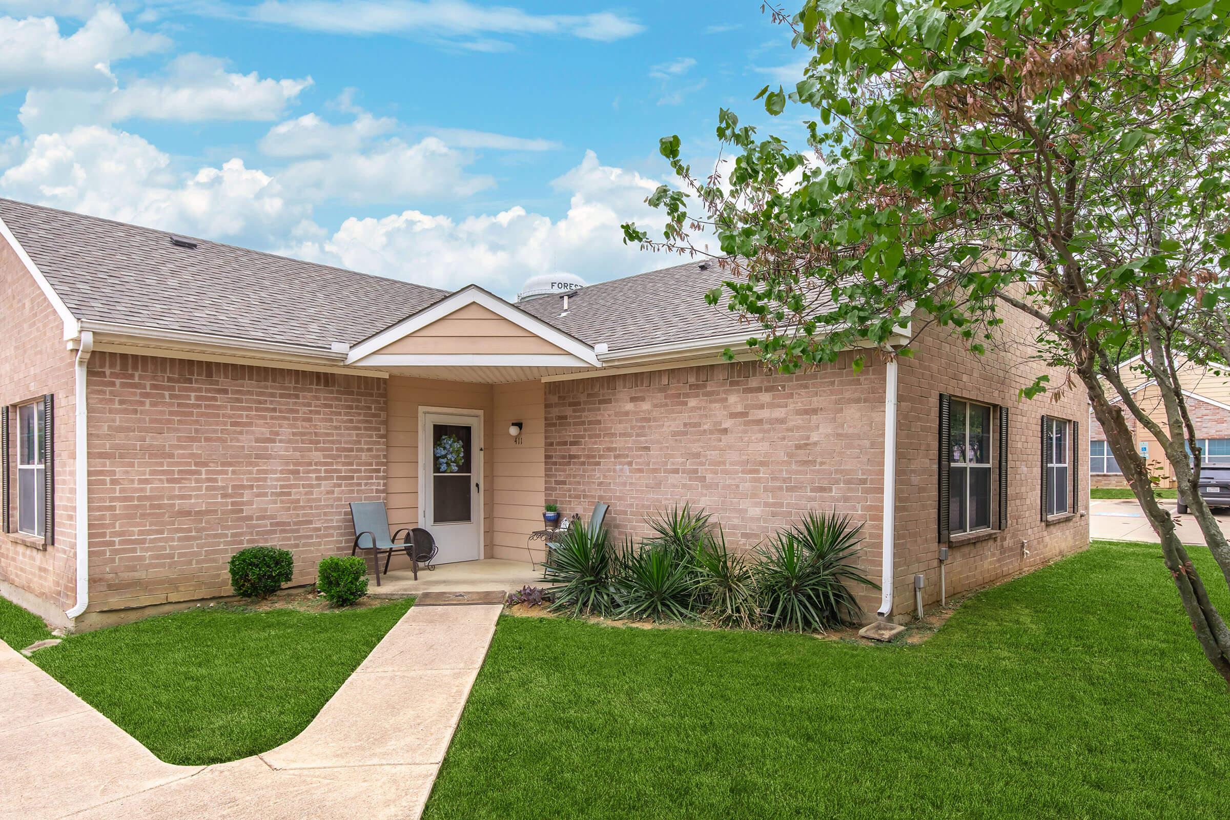 A well-maintained single-story brick home with a spacious front yard. The entrance features a small porch with a chair, surrounded by green grass and decorative shrubs. The sky is clear with some clouds, enhancing the inviting atmosphere of the property.