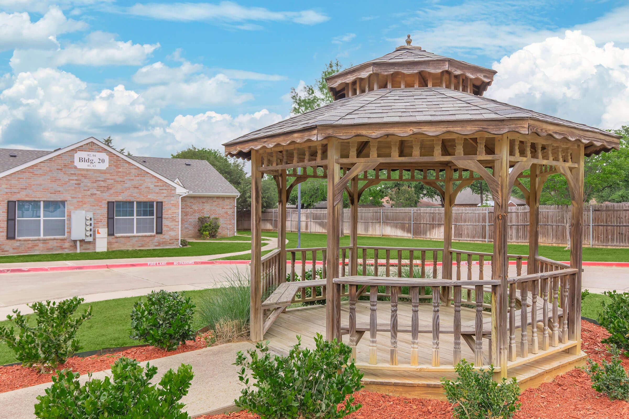 A wooden gazebo with a shingled roof sits in a well-maintained outdoor area, surrounded by small shrubs and mulch. In the background, there is a brick building with a sign, and a clear blue sky with scattered clouds overhead. The scene conveys a peaceful, inviting setting.