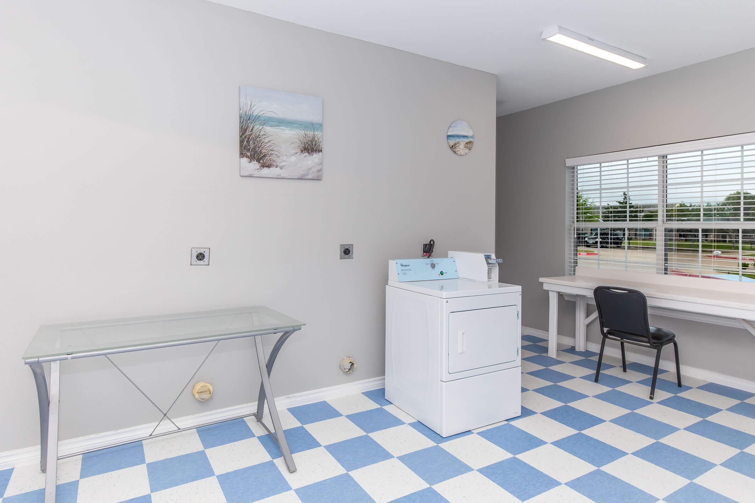 A bright laundry room featuring a white washing machine, a glass-top table, and a black chair. The walls are painted light gray, and there's a window with blinds allowing natural light. A coastal-themed painting hangs on the wall, along with a decorative item, and the floor has a blue and white checkered pattern.