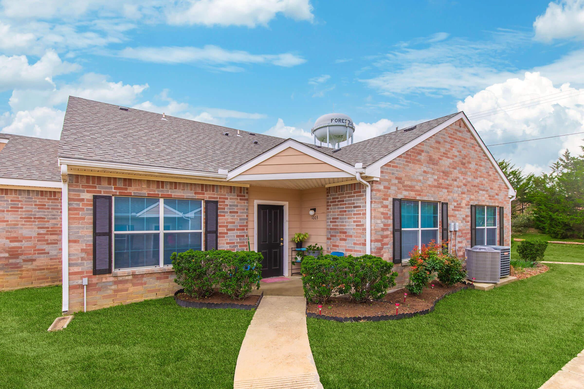 A brick bungalow with a well-maintained lawn and colorful flower beds. The house features a welcoming front porch, a black front door, and several windows with black shutters. In the background, a water tower is visible against a blue sky with fluffy clouds.