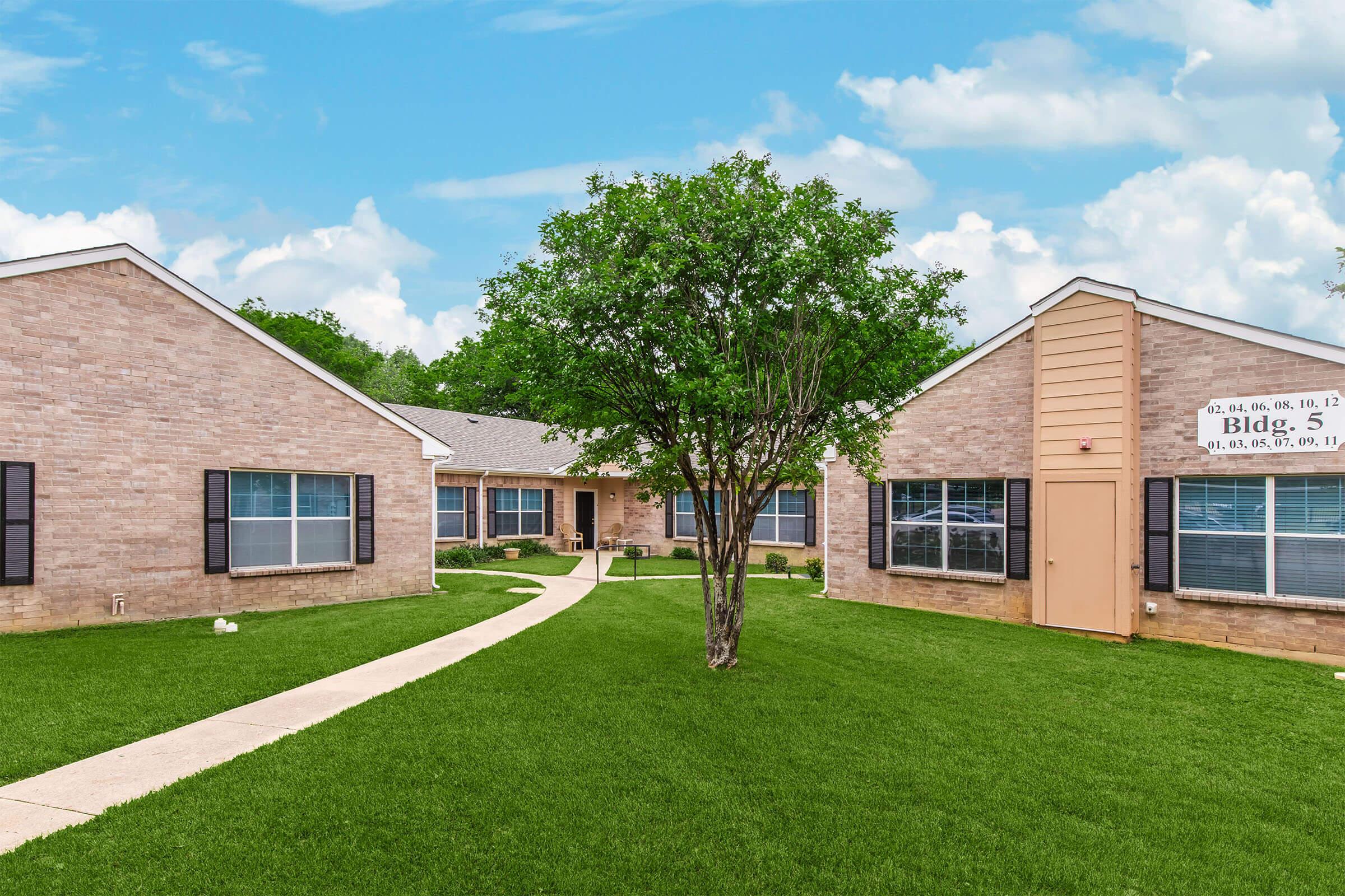 A well-maintained brick apartment complex featuring two buildings with large windows, a green lawn, and a tree in the center. A paved pathway leads through the grassy area, providing access to the entrances of the buildings. The sky is clear with fluffy clouds.
