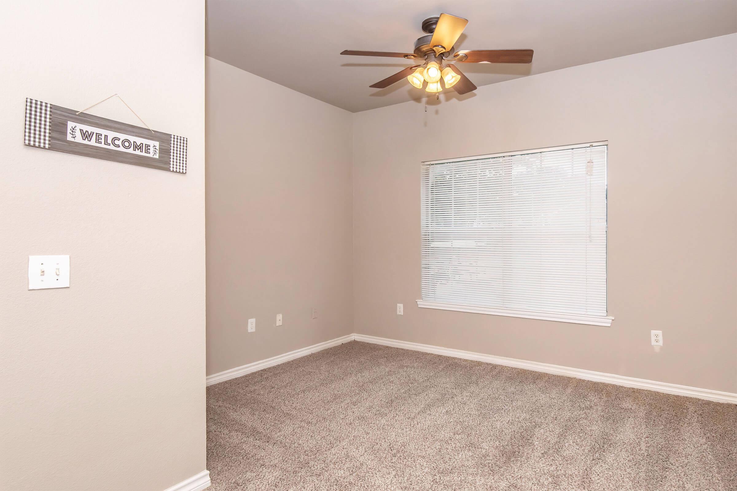 A cozy room featuring light-colored walls and a ceiling fan with light fixtures. The space has a window with blinds allowing natural light, and plush, carpeted flooring. A "Welcome" sign hangs on the wall, creating a warm, inviting atmosphere.
