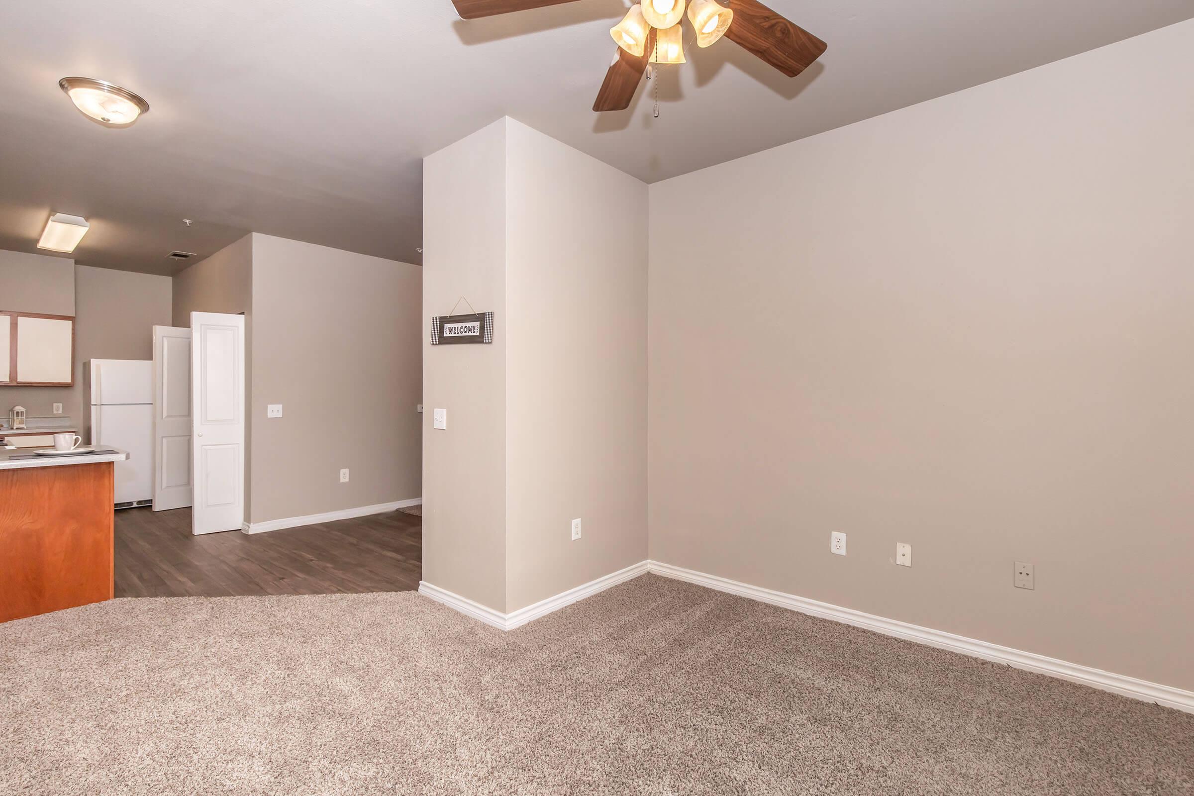 A spacious, empty living area with light-colored walls and beige carpet. A ceiling fan is visible, and there's a doorway leading to a kitchen area in the background. The interior appears modern and well-lit, with no furniture present.