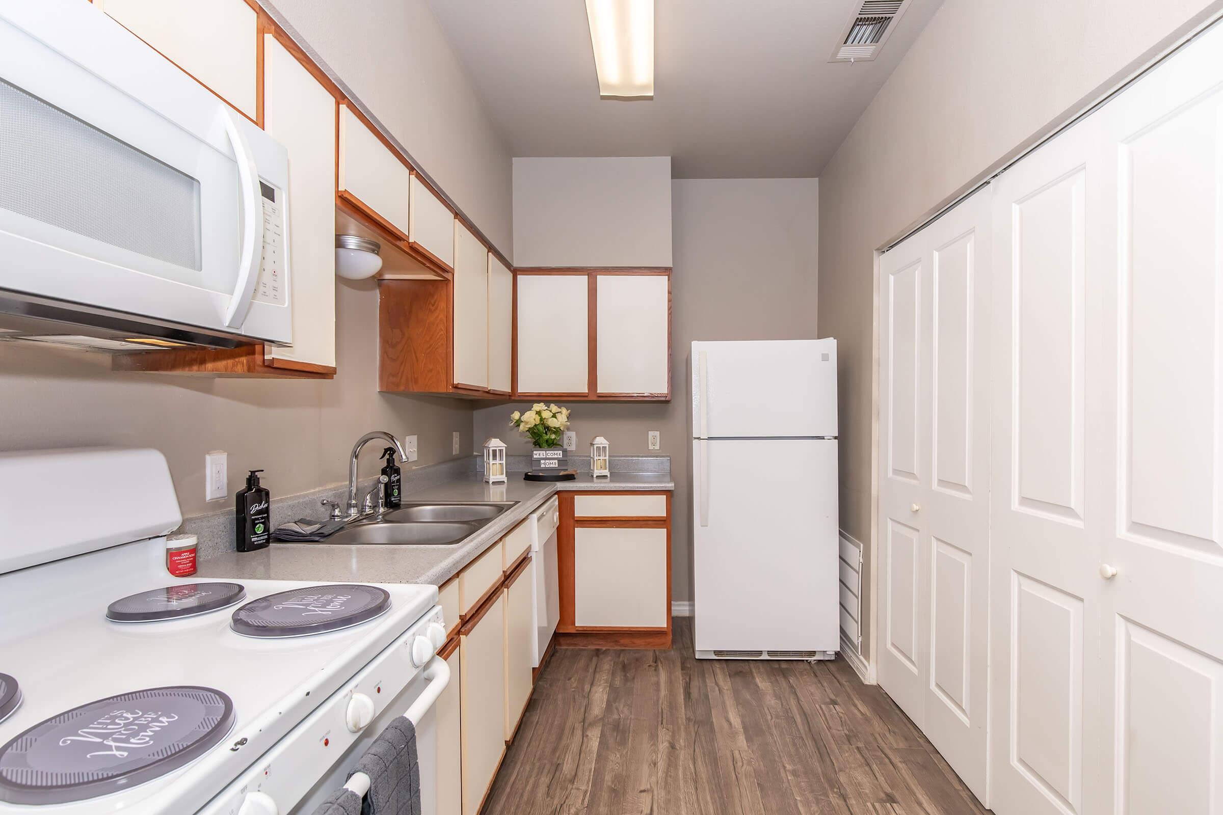 A modern kitchen featuring light wood cabinets, a white refrigerator, a sink with a faucet, and a stove. Countertops are clean and clutter-free. The room has light-colored walls and vinyl flooring, with a small arrangement of flowers on the counter. There are closed double doors leading to a storage area.