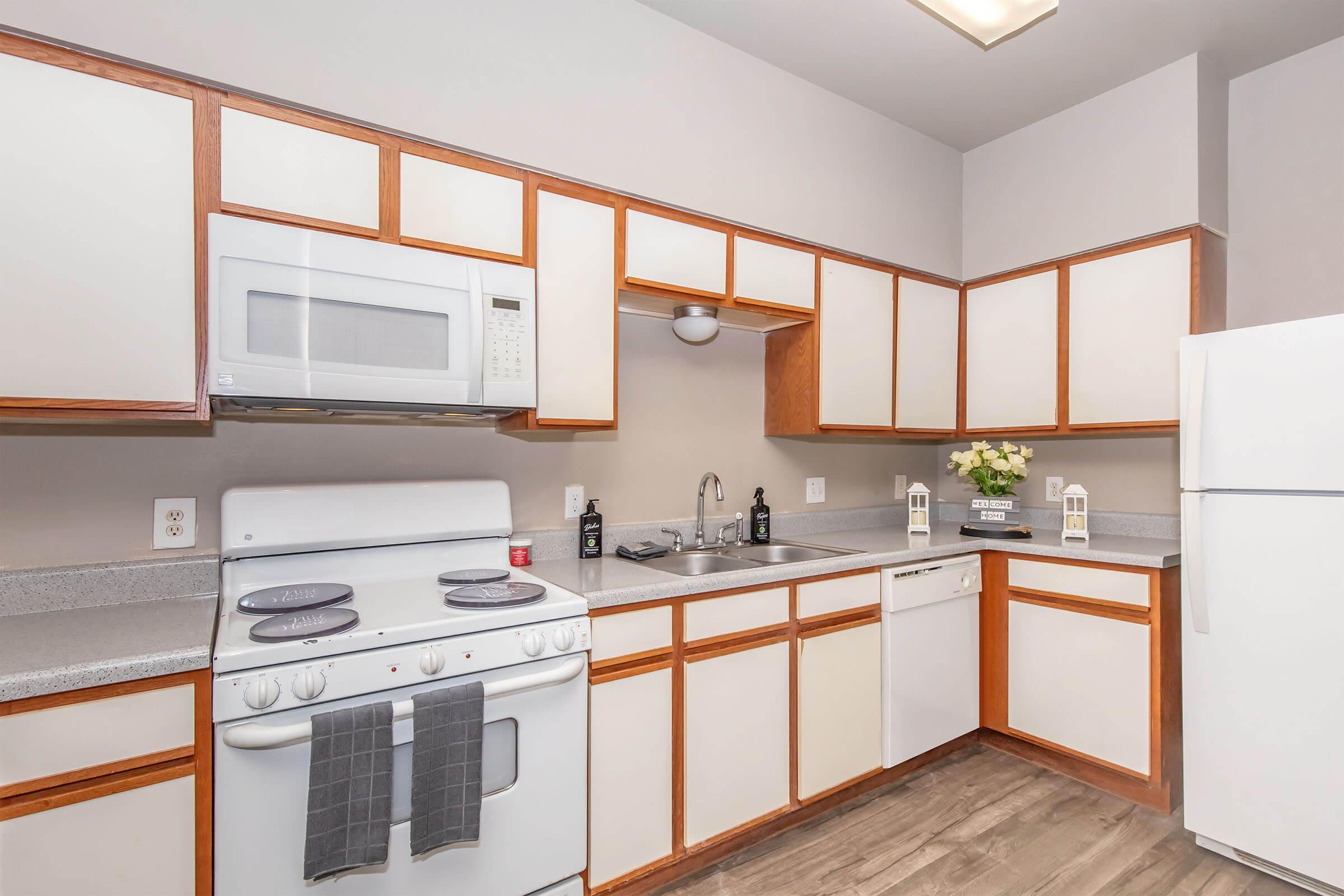 A modern kitchen featuring light wood cabinets, a white microwave, stove, and refrigerator. The countertop is grey, with a sink and dishwasher. Decorative elements include a flower arrangement and small lanterns. The walls are painted a soft color, creating a bright and inviting atmosphere.