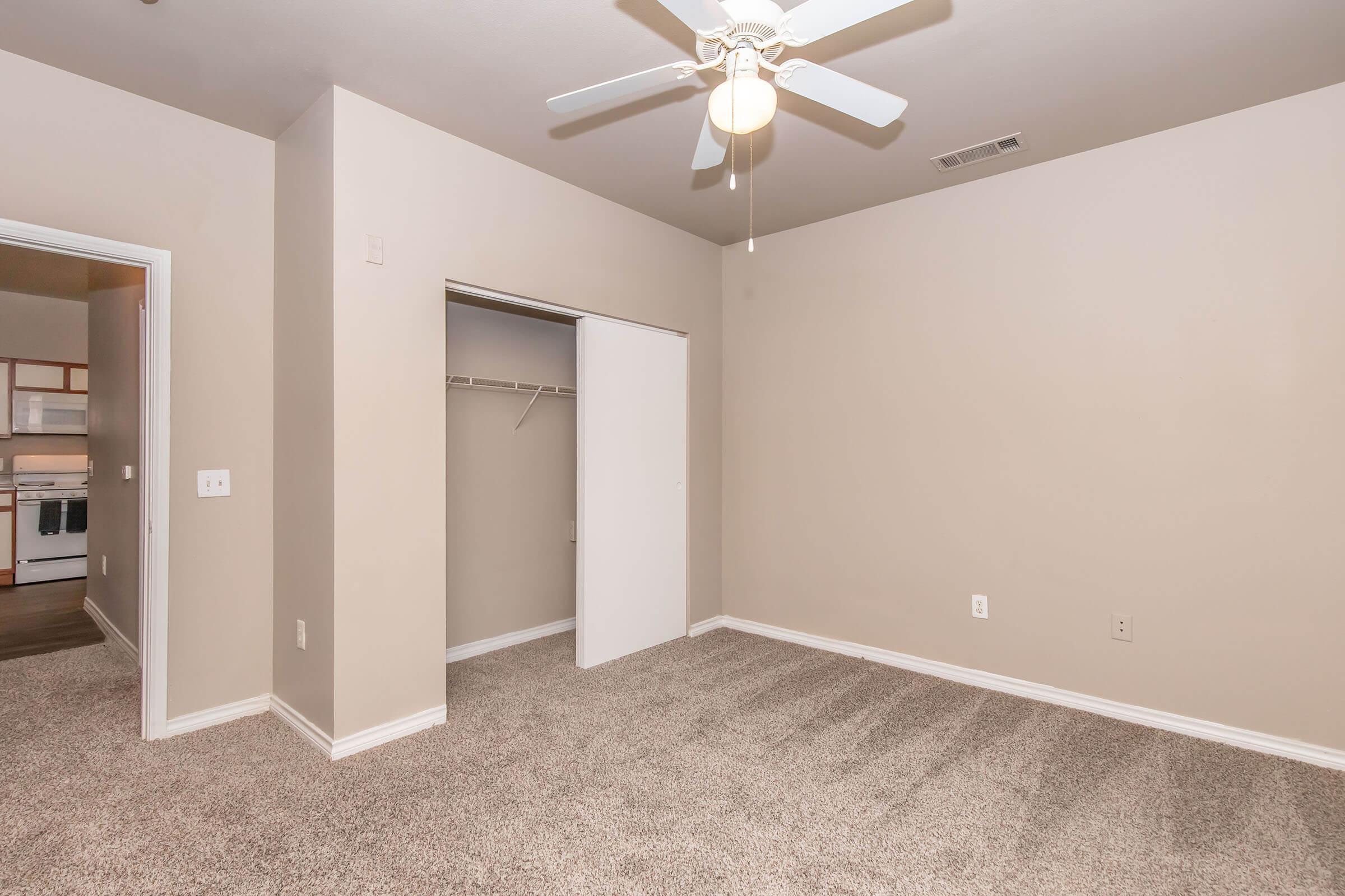 A vacant room featuring light beige walls and carpeted flooring. There is a ceiling fan with light fixtures and a closet with sliding doors. The doorway to the left leads to a kitchen area, partially visible. The overall space is well-lit and ready for customization.