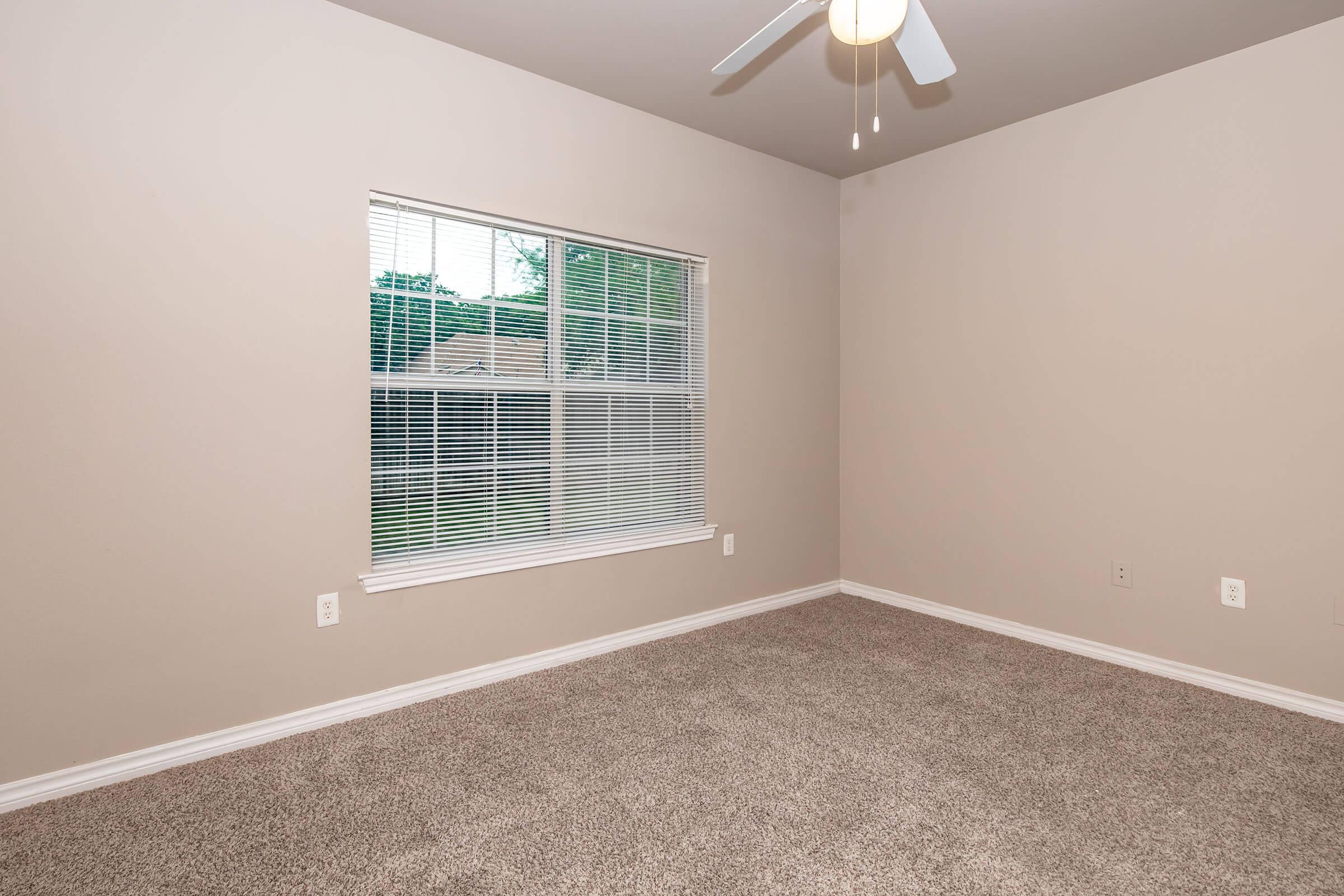 A vacant room with light beige walls and carpeted flooring. The room features a ceiling fan and a large window with white blinds that allows natural light in, revealing greenery outside. There are no furnishings or decorations in the space, creating a clean and minimalistic appearance.