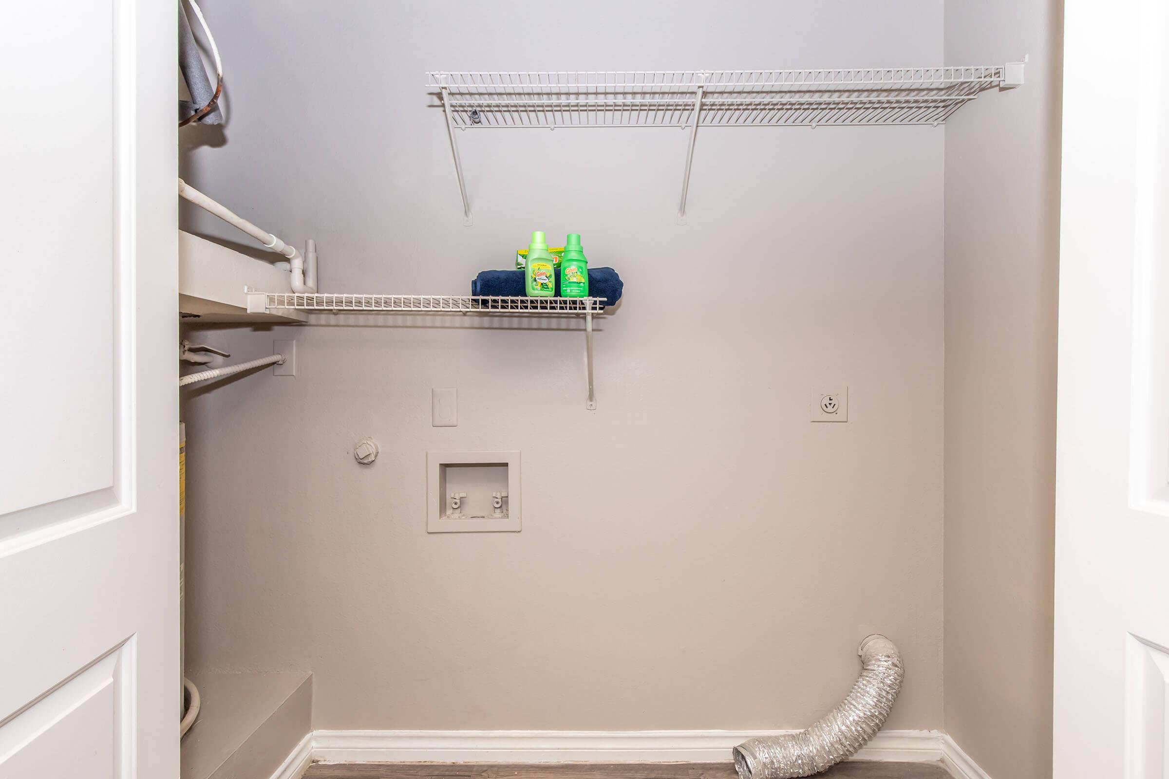 A small, empty laundry room with two wire shelves. One shelf holds two green bottles, while the other is empty. There's a dryer vent hose on the floor. The walls are painted light gray, and the flooring appears to be laminate. The room has a minimalist and organized look.