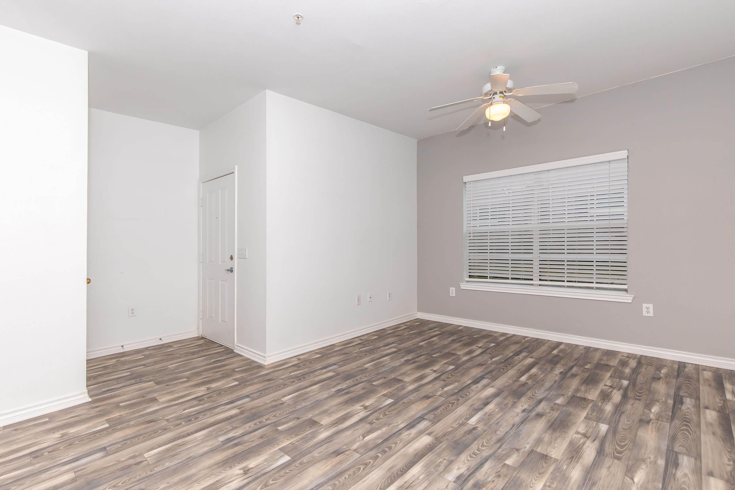 A spacious, empty room with light gray walls and a ceiling fan. The floor features modern laminate flooring in wood tones. A large window with white blinds allows natural light, and there's a closed door visible on the left side of the room.