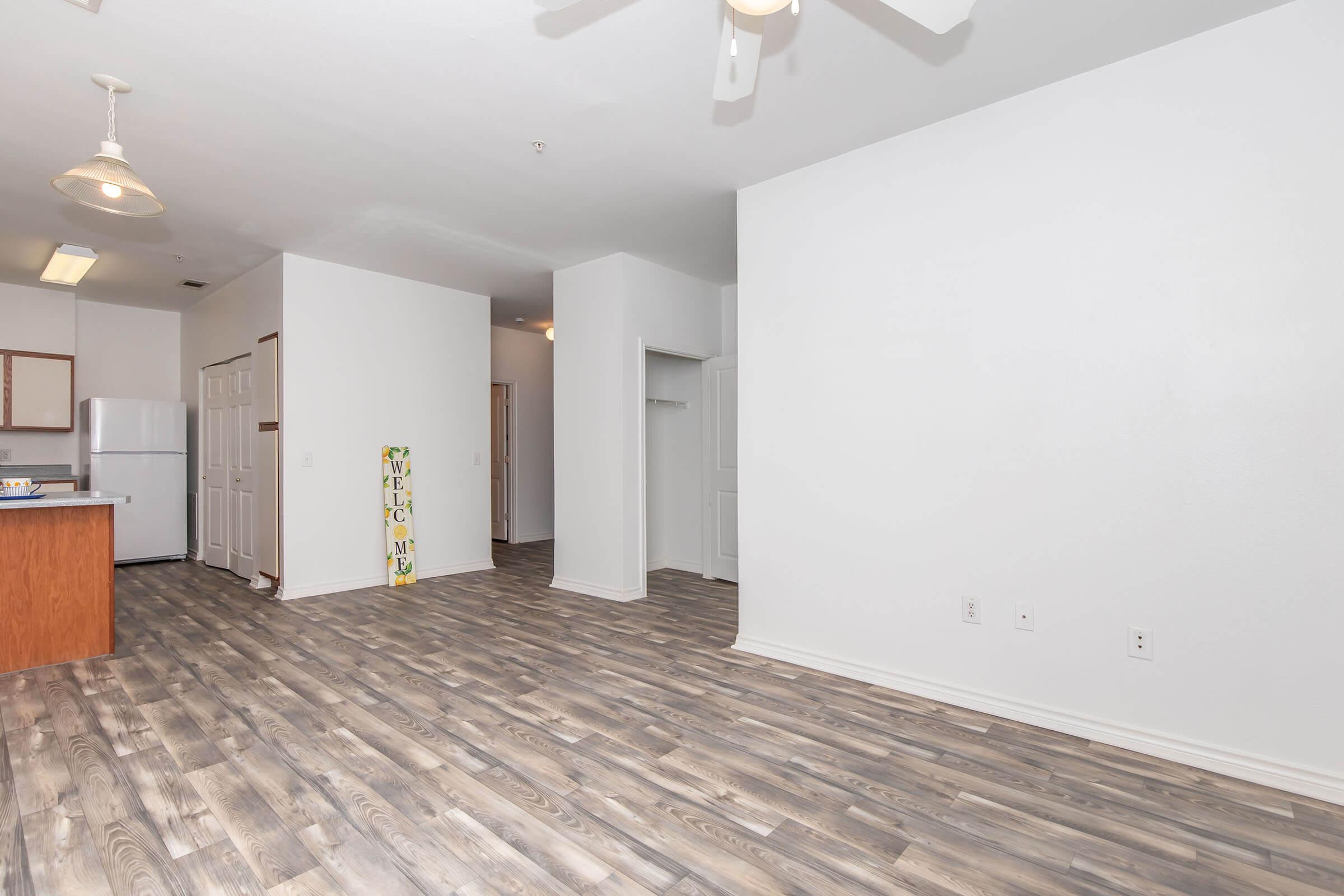 A spacious, well-lit living area features light-colored walls and a modern wooden floor. A ceiling fan hangs above, and an open kitchen with a refrigerator is visible to the left. There is a welcome sign on the floor, with a doorway leading to another room seen in the background. The overall atmosphere is bright and inviting.