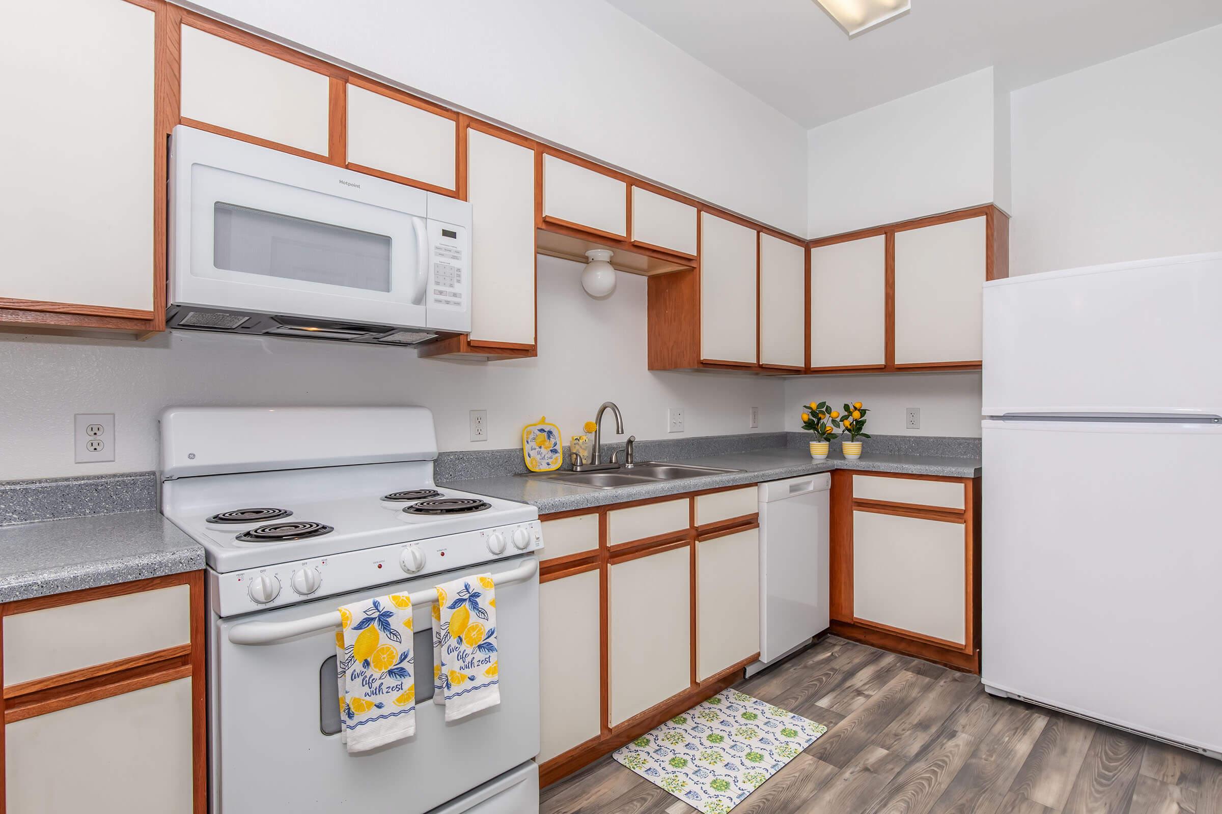A bright, modern kitchen featuring white and wooden cabinetry, a white stove and microwave, and a refrigerator. The countertop is gray, and there’s a small sink with a vase of yellow flowers nearby. A colorful kitchen mat adds a pop of color to the floor. Natural light illuminates the space.