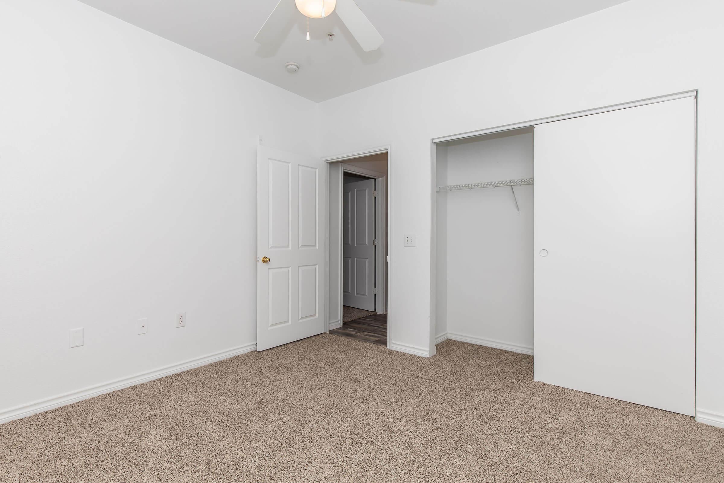 Empty room with white walls and a ceiling fan. The floor is covered with light brown carpet. There are two doorways: one leads to another room and the other has a sliding closet door. Natural light enters through an unseen window, creating a bright atmosphere.