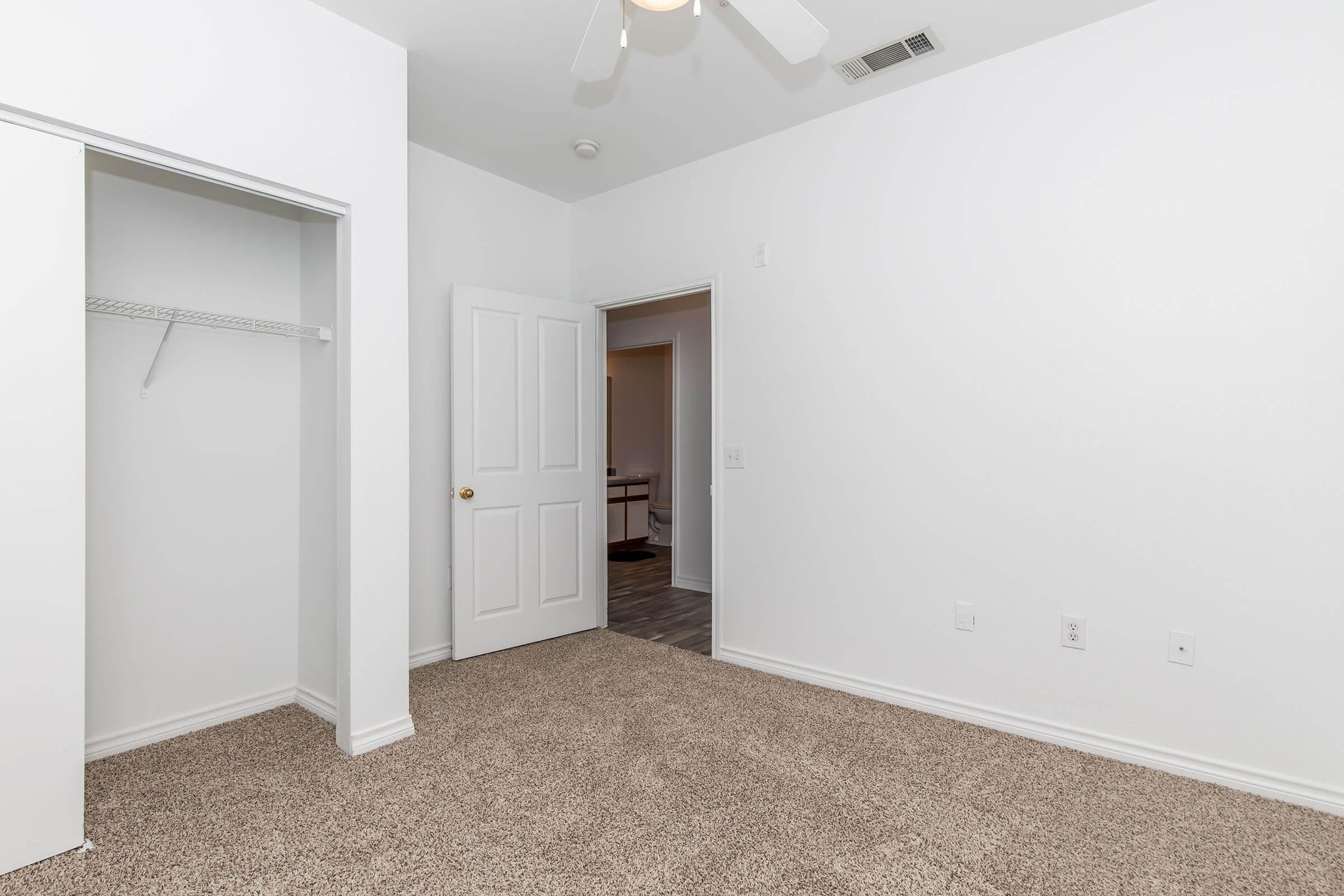 A bright, empty room featuring beige carpet flooring, white walls, and a ceiling fan. There is an open closet space with a wire shelf on the left, and a closed door leading to another area in the background. The overall atmosphere is spacious and clean.