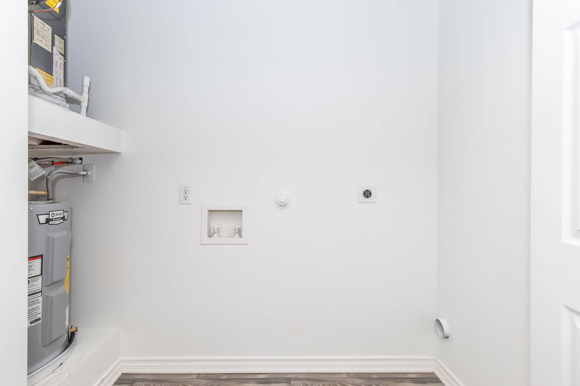 A small laundry room with white walls and hardwood-style flooring. The space includes a water heater on the left and a wall with two openings, one circular and one square, for plumbing. The area appears clean and minimalistic, with no visible appliances or furniture.