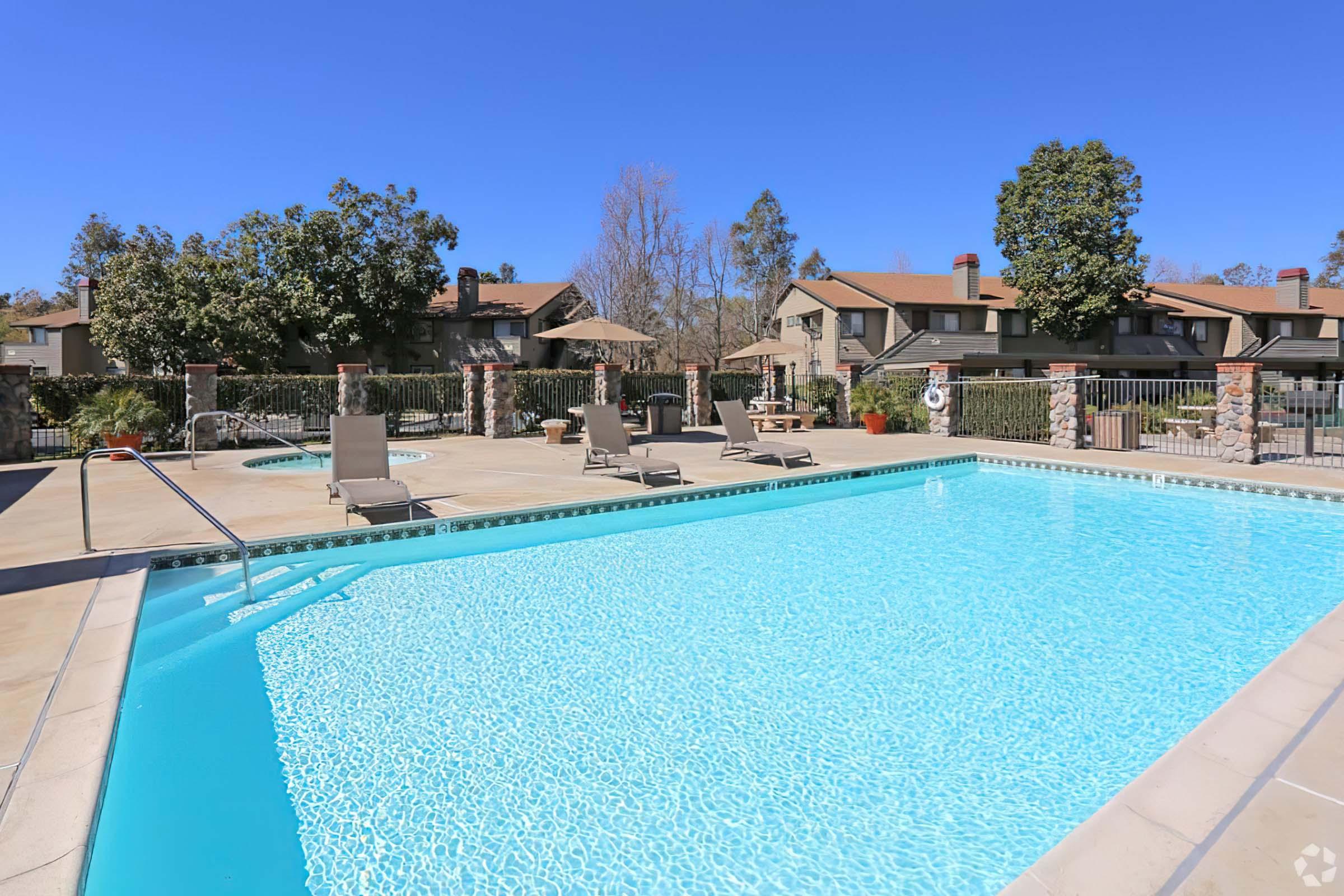 A bright outdoor swimming pool surrounded by lounge chairs, with clear blue water reflecting the sky. In the background, there are trees and residential buildings, indicating a pleasant community setting. The sun is shining, creating a warm and inviting atmosphere.