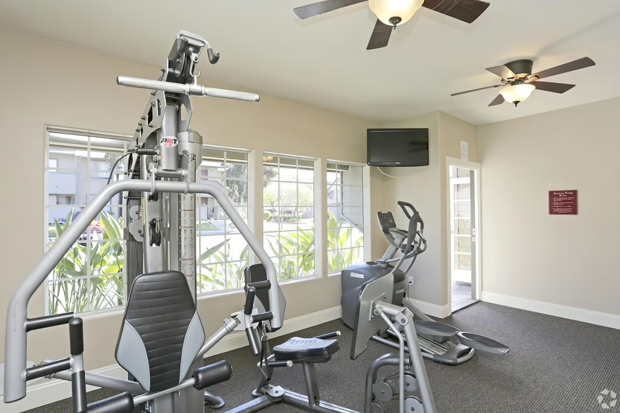 A well-lit gym with exercise equipment, including a multi-gym machine and an elliptical trainer. Two ceiling fans are visible, and there are large windows allowing natural light. A wall-mounted TV is also present, enhancing the workout environment.