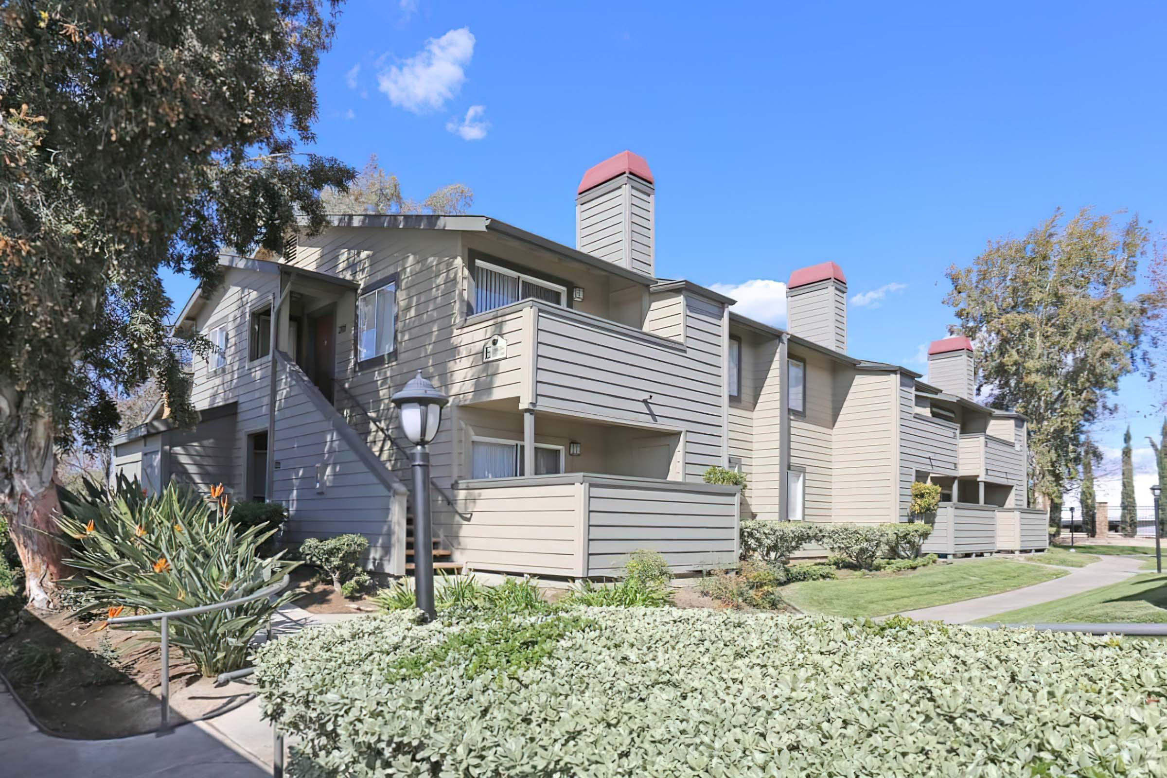 A modern apartment complex featuring multiple two-story buildings with balconies, surrounded by green landscaping and trees under a blue sky. The design includes red-topped chimneys and a pathway leading to the entrance.