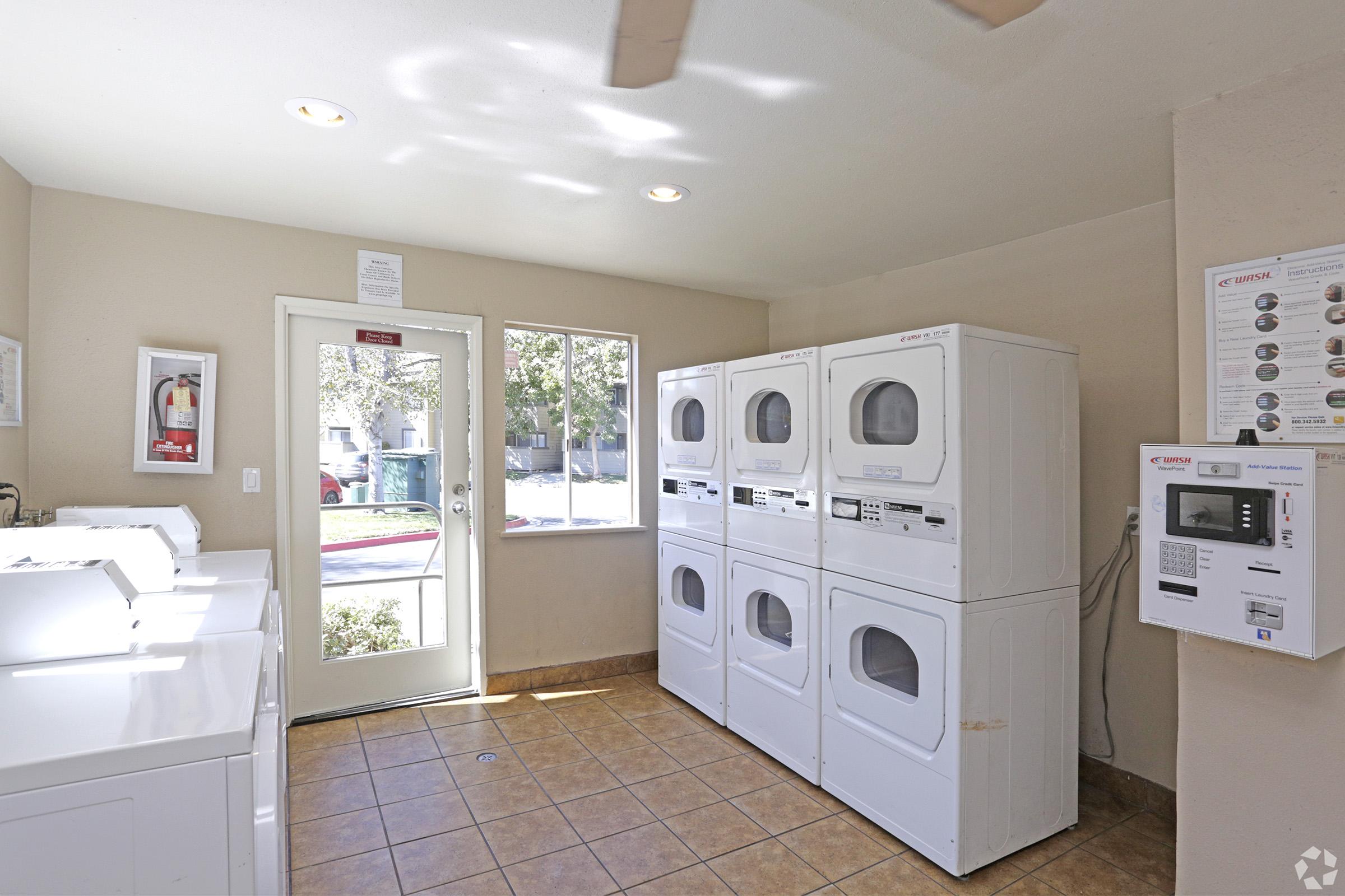 A well-lit laundry room featuring multiple white washing machines and dryers arranged against a wall. There is a large window allowing natural light in, a door leading outside, and a payment machine mounted on the wall. The tiled floor adds a clean and organized appearance to the space.