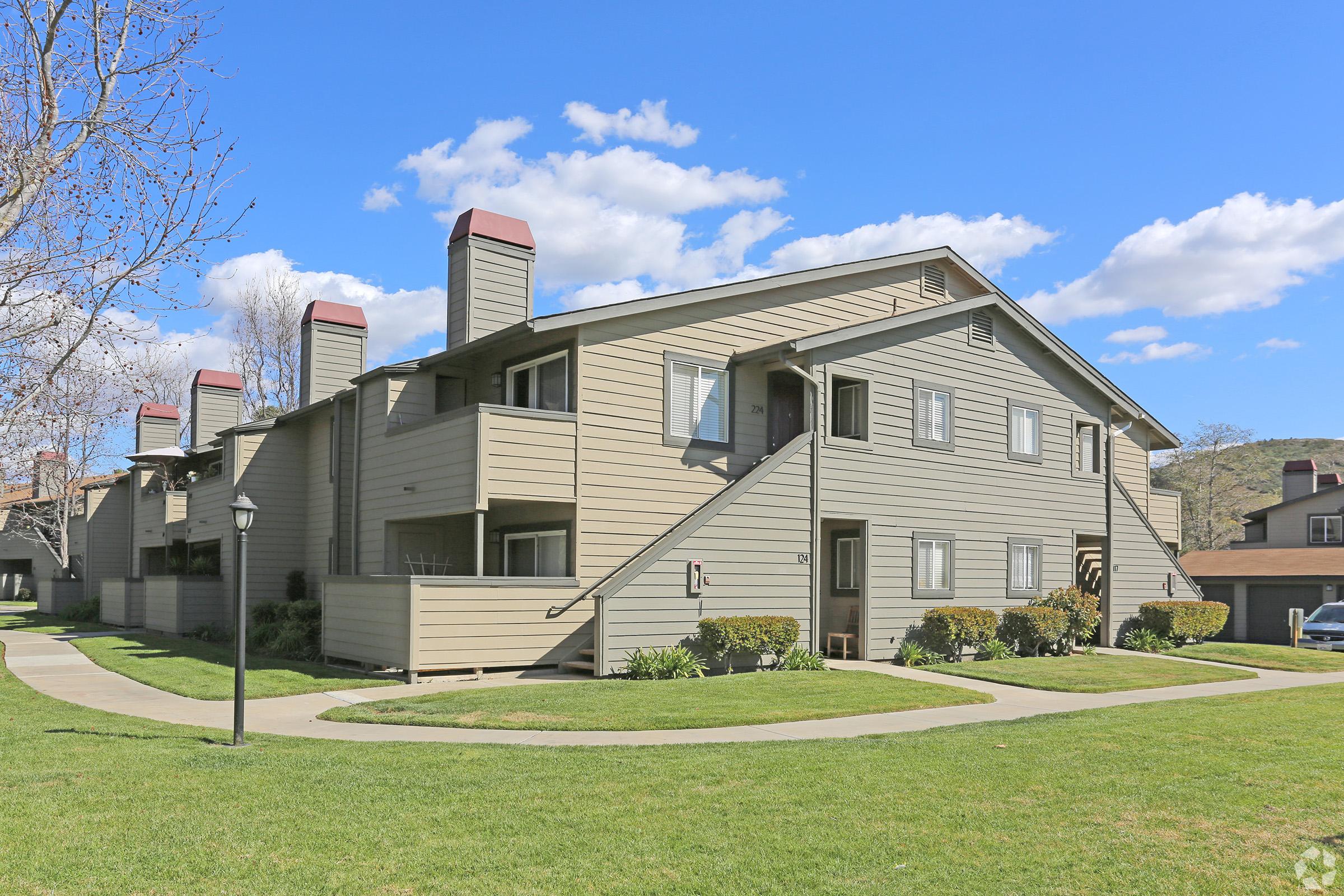 A two-story residential building with a light brown wooden exterior, featuring multiple balconies and chimneys. The building is surrounded by green grass and small shrubs, with a pathway leading to the entrance. The sky is clear with a few clouds, creating a bright atmosphere.