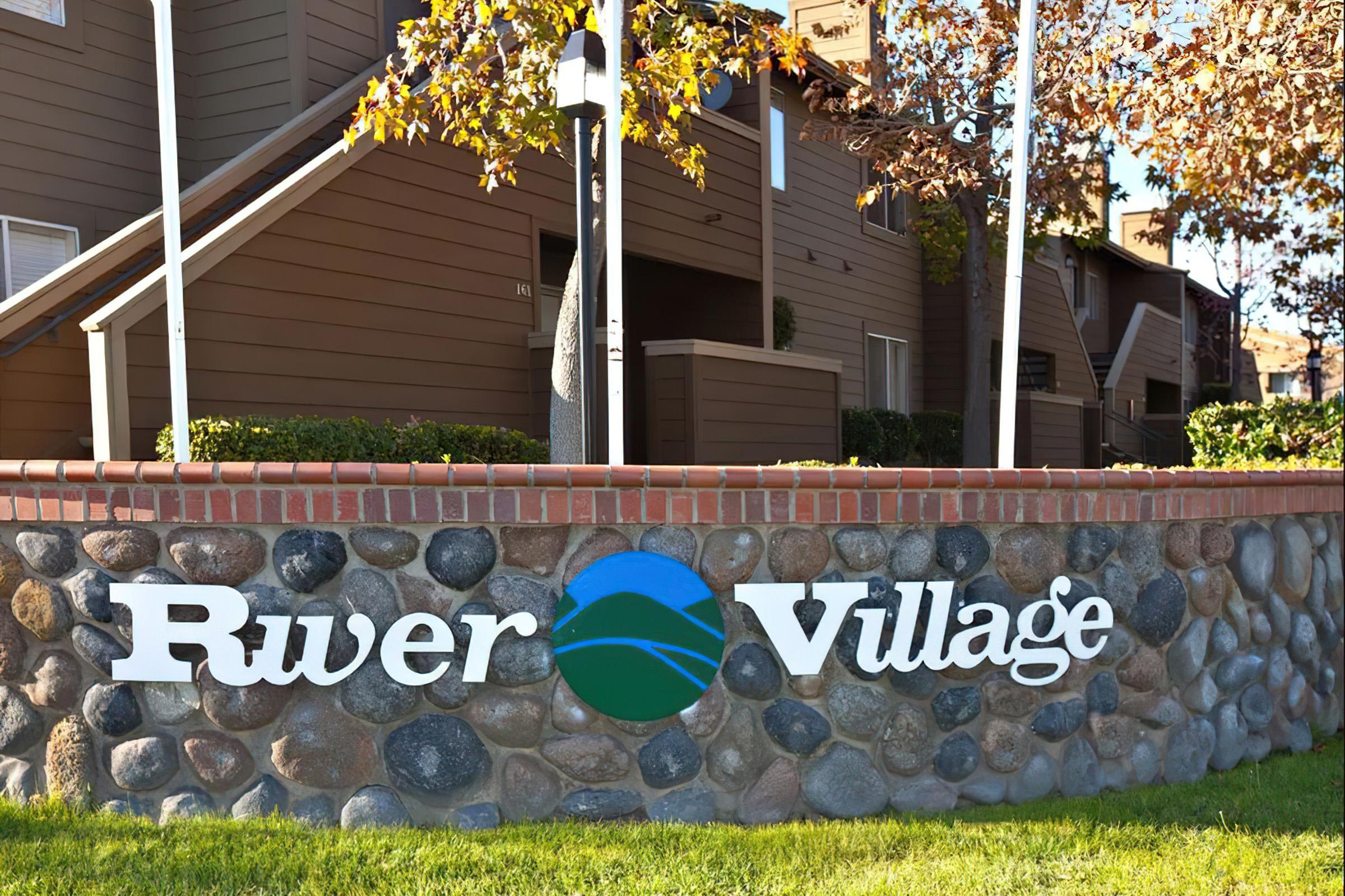 Sign displaying "River Village" with a blue and green logo, situated in front of a stone wall. The background features residential buildings and trees with autumn leaves, suggesting a cozy community atmosphere. The lower grass area is neatly maintained.