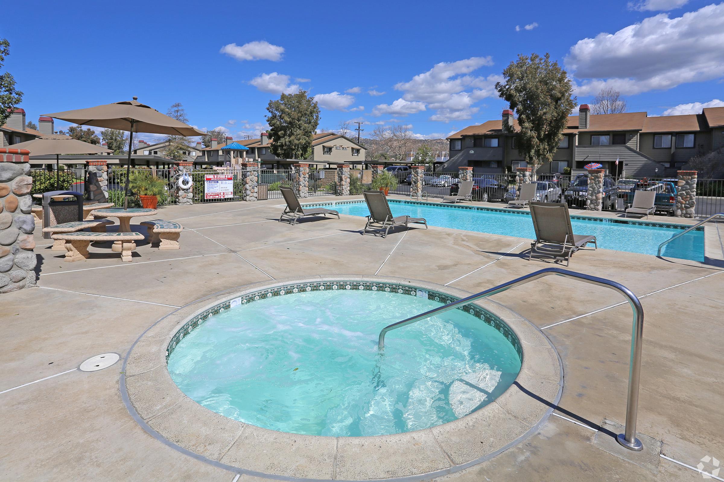 A hot tub filled with bubbling water next to a swimming pool in a residential area. Lounge chairs are arranged around the pool, and there are patio umbrellas providing shade. In the background, there are buildings surrounded by trees under a blue sky with fluffy clouds.