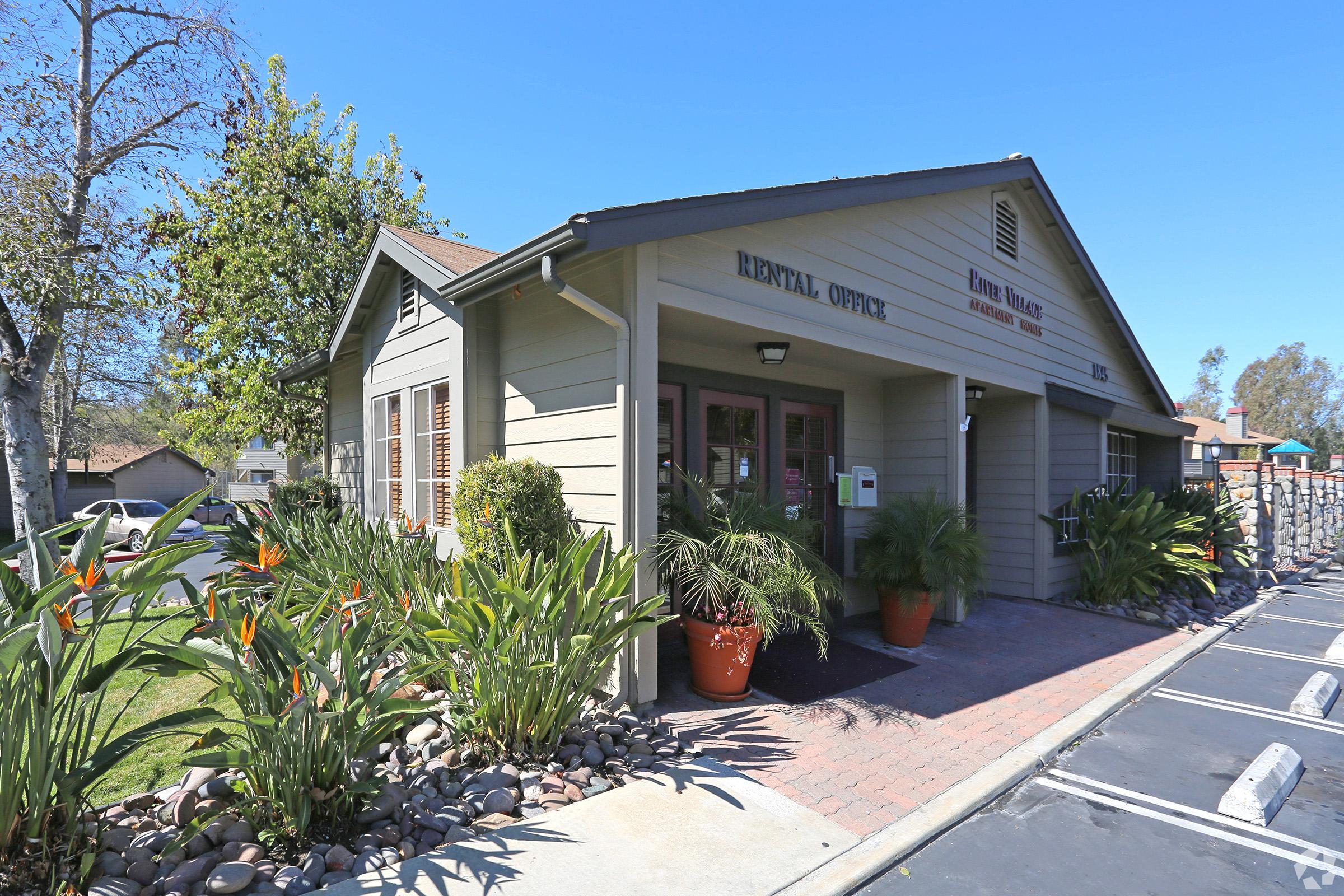 A one-story dental office with a welcoming entrance. The building features a covered porch, large windows, and a sign that reads "Dental Office." Lush greenery and potted plants surround the entrance, adding a friendly touch. A parking lot is visible in front, with clear blue skies above.