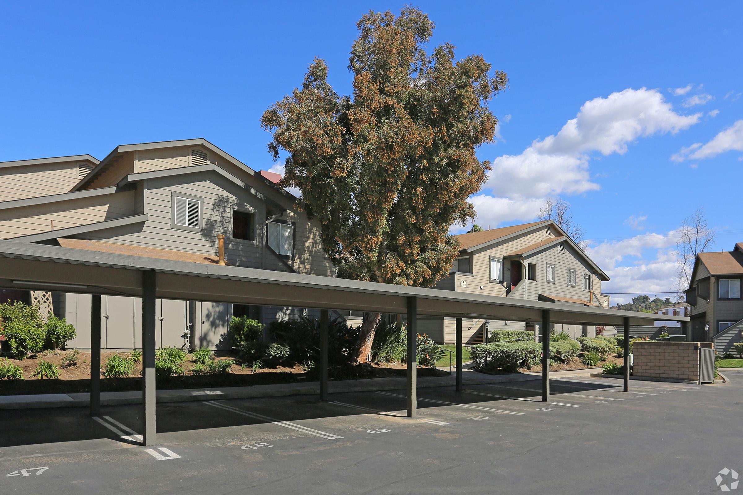 A peaceful residential area featuring several multi-unit buildings, a large tree, and covered parking spaces. The sky is clear with a few clouds, and the landscaping is well-maintained, enhancing the property's appeal.