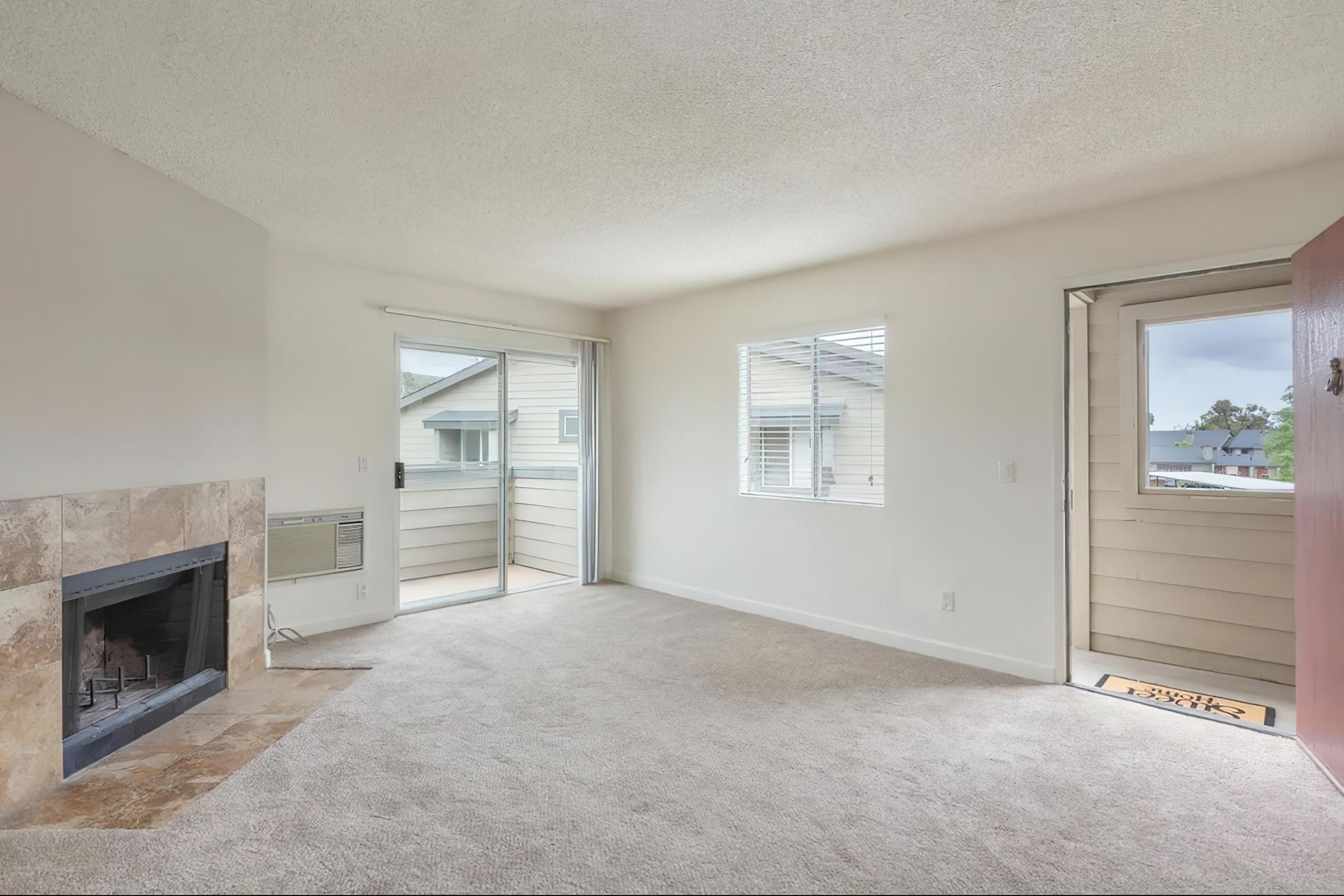 A spacious, well-lit living room with light-colored carpet and white walls. Features include a stone fireplace, sliding glass doors leading to a balcony, and a front door with a welcome mat. Windows provide natural light, and there is an air conditioning unit on the wall.