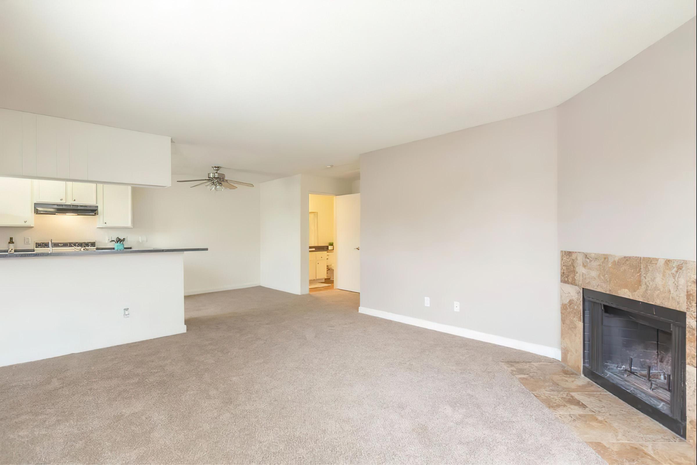 A spacious, empty living room with light-colored carpet and walls. There is a ceiling fan and a fireplace made of stone. The kitchen is partially visible in the background, featuring light cabinetry. The overall atmosphere is bright and open, creating a welcoming feeling.