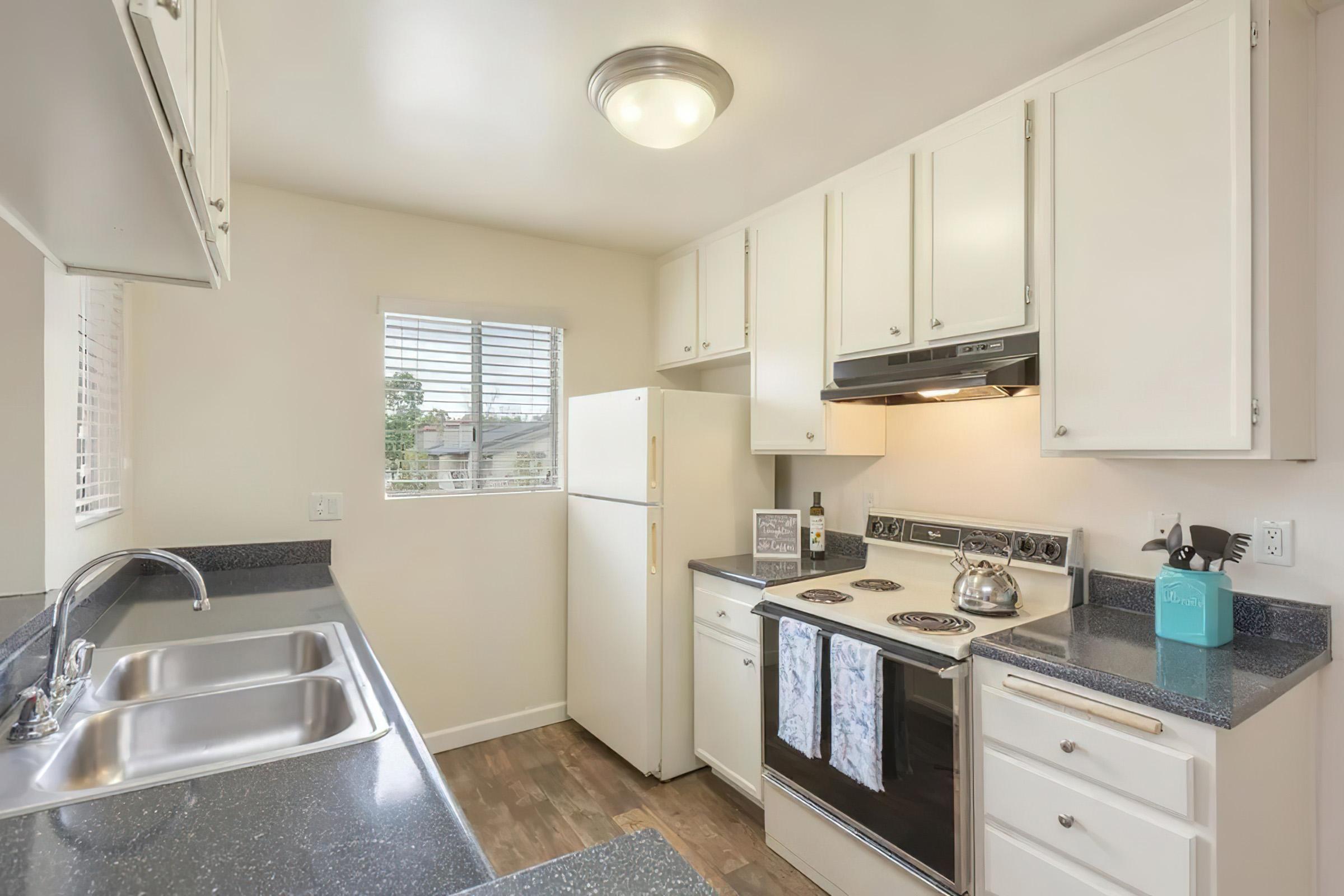 A bright and modern kitchen featuring white cabinetry, a standard refrigerator, an oven with a stovetop, and a double-basin sink. The countertop is dark and smooth, complementing the appliances. Natural light streams in through a window with white blinds, enhancing the inviting ambiance.