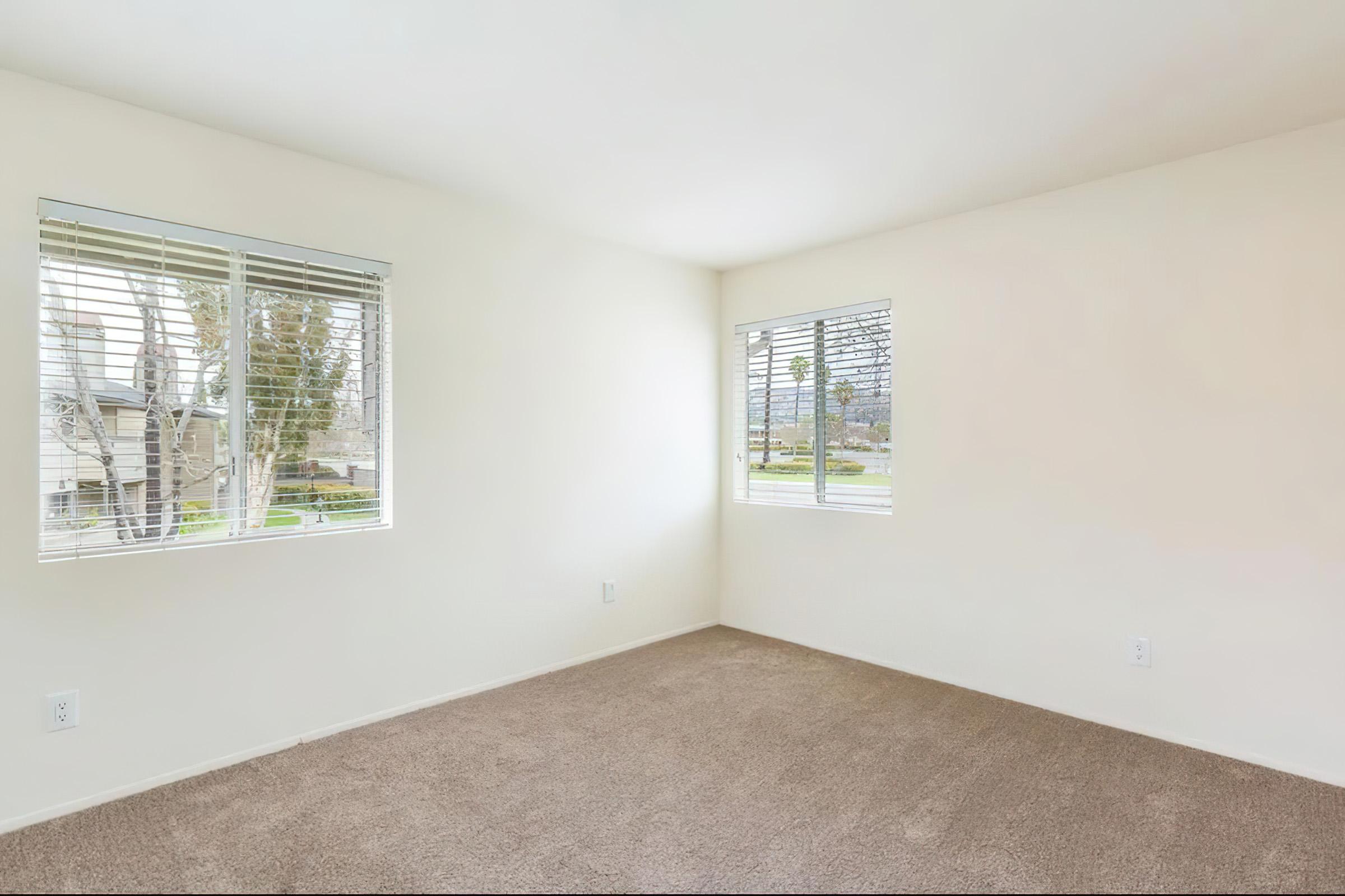 Empty room featuring light-colored walls, two windows with blinds, and carpeted flooring. Natural light streams in, illuminating the space, which has a minimalist design. The view outside the windows includes greenery and possibly a residential area.